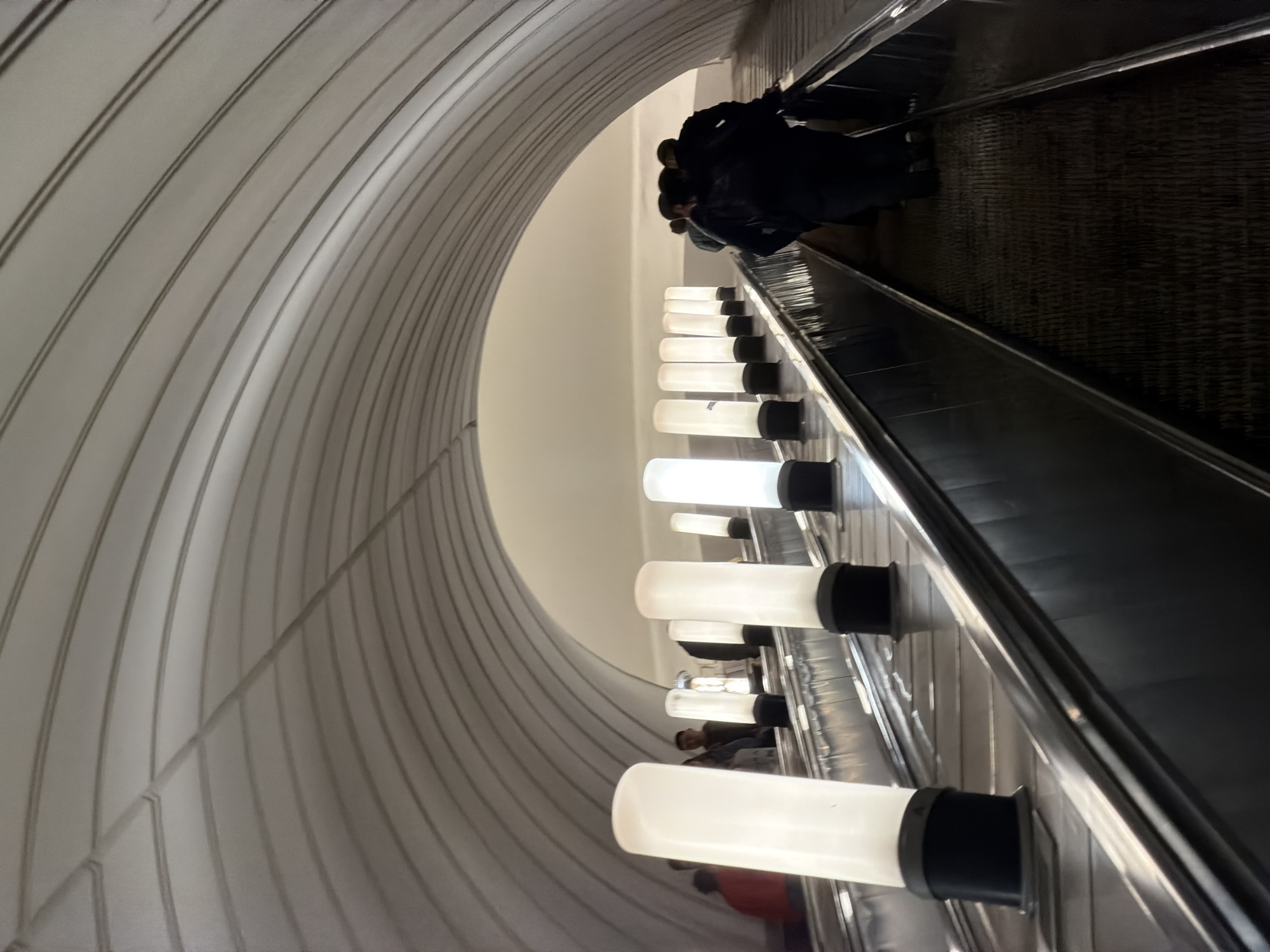The image shows an escalator tunnel in the Moscow Metro, Russia. The tunnel has a curved, white-paneled wall on the left and a row of cylindrical lights mounted along the escalator's handrail. People are riding the escalator, moving upwards. The escalator steps are visible, and the handrail is made of metal.