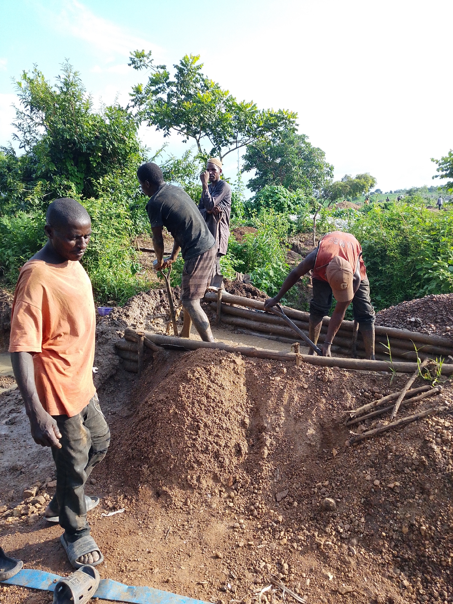 The image depicts a scene of manual labor in what appears to be an open-pit mine or construction site in Otulu, Nigeria. Four individuals are actively engaged in work. In the foreground, a man wearing an orange t-shirt and dark pants, and sandals, stands facing left, looking down. To his right and slightly behind him, another man in a dark t-shirt and plaid shorts is using a shovel to dig into a pile of dirt. Further back, a third individual, wearing a patterned garment and a cap, appears to be observing. In the mid-ground, a fourth person, dressed in an orange sleeveless shirt and dark pants, is bent over, using a tool, possibly a shovel or pickaxe, to work in a trench lined with wooden planks. The environment is characterized by rough terrain with piles of soil and rocks, and lush green vegetation in the background. The ground is muddy in some areas, suggesting recent rain or water presence. The sky is visible in the upper portion of the image, with some clouds. The overall impression is one of strenuous physical work in a rural or undeveloped setting.