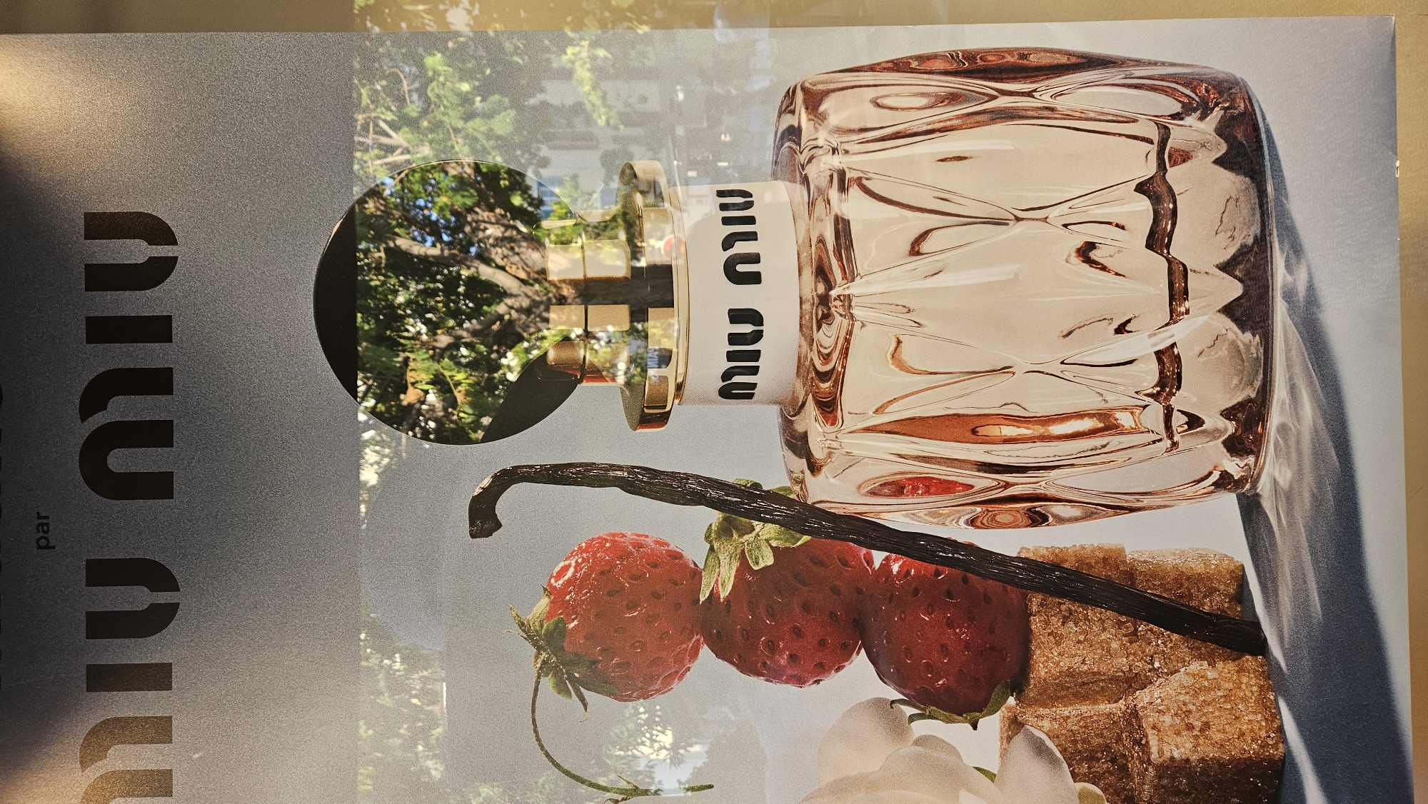 A close-up studio shot showcases a perfume bottle with a textured, rose-gold colored glass body. The bottle has a white label with the "MIU MIU" logo in black lettering. The cap is gold and cylindrical, with a circular opening reflecting trees and foliage. To the left of the bottle are three red strawberries stacked vertically, a vanilla bean, and two cubes of brown sugar. The background is a gradient of gray, with a hint of the reflection of an outdoor environment, possibly a city street or park, suggesting the image was taken in L'Île-Rousse, France, perhaps in a shop window display. The lighting creates sharp shadows and highlights on the perfume bottle and the surrounding objects.