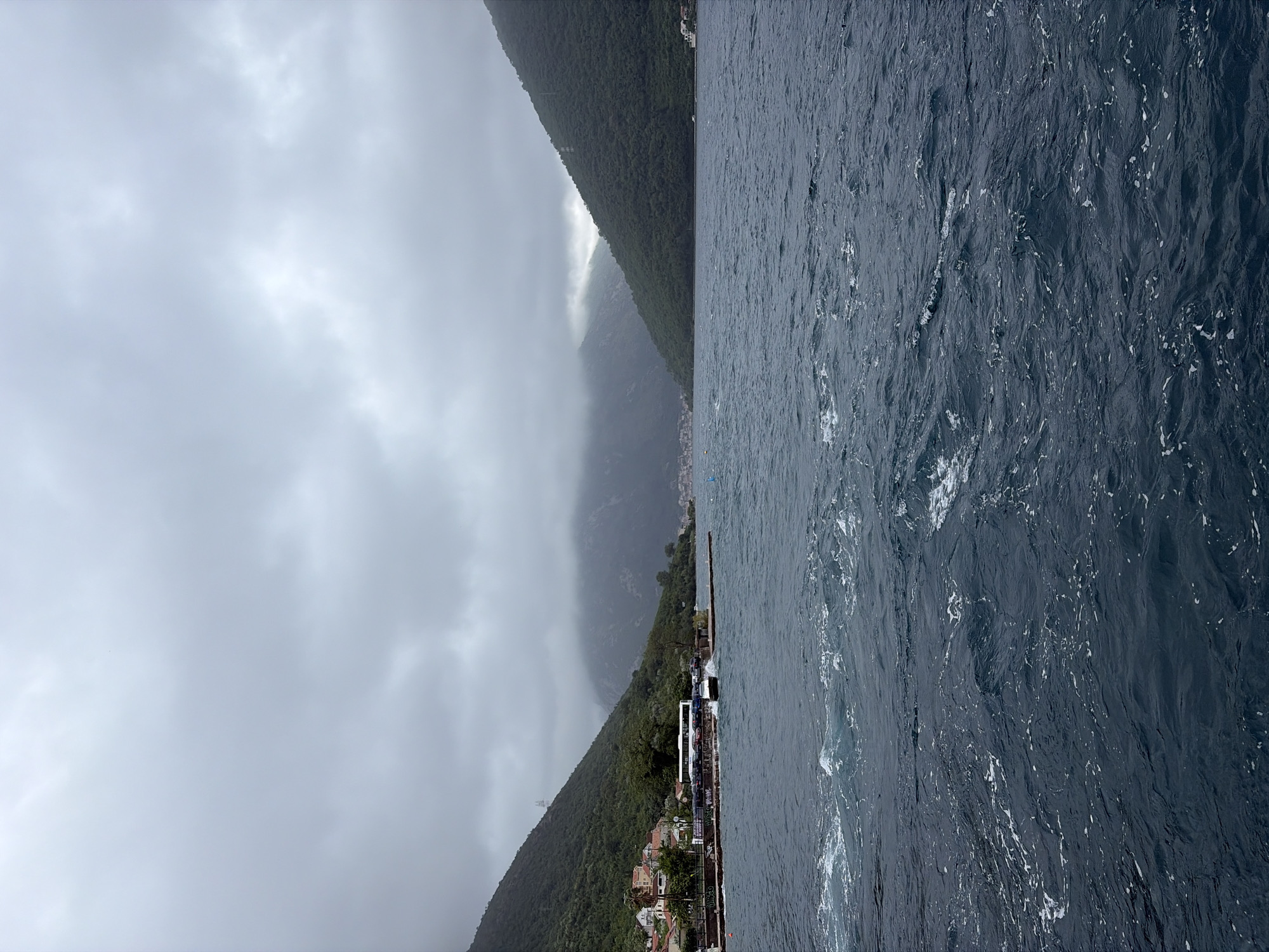 The image depicts a view in Đurići, Montenegro. The left side of the frame is dominated by a cloudy sky and forested mountains partially obscured by mist. Buildings are visible at the base of the mountain along the waterfront. A pier extending into the water is visible in the lower left. The right side of the image shows dark, choppy water.