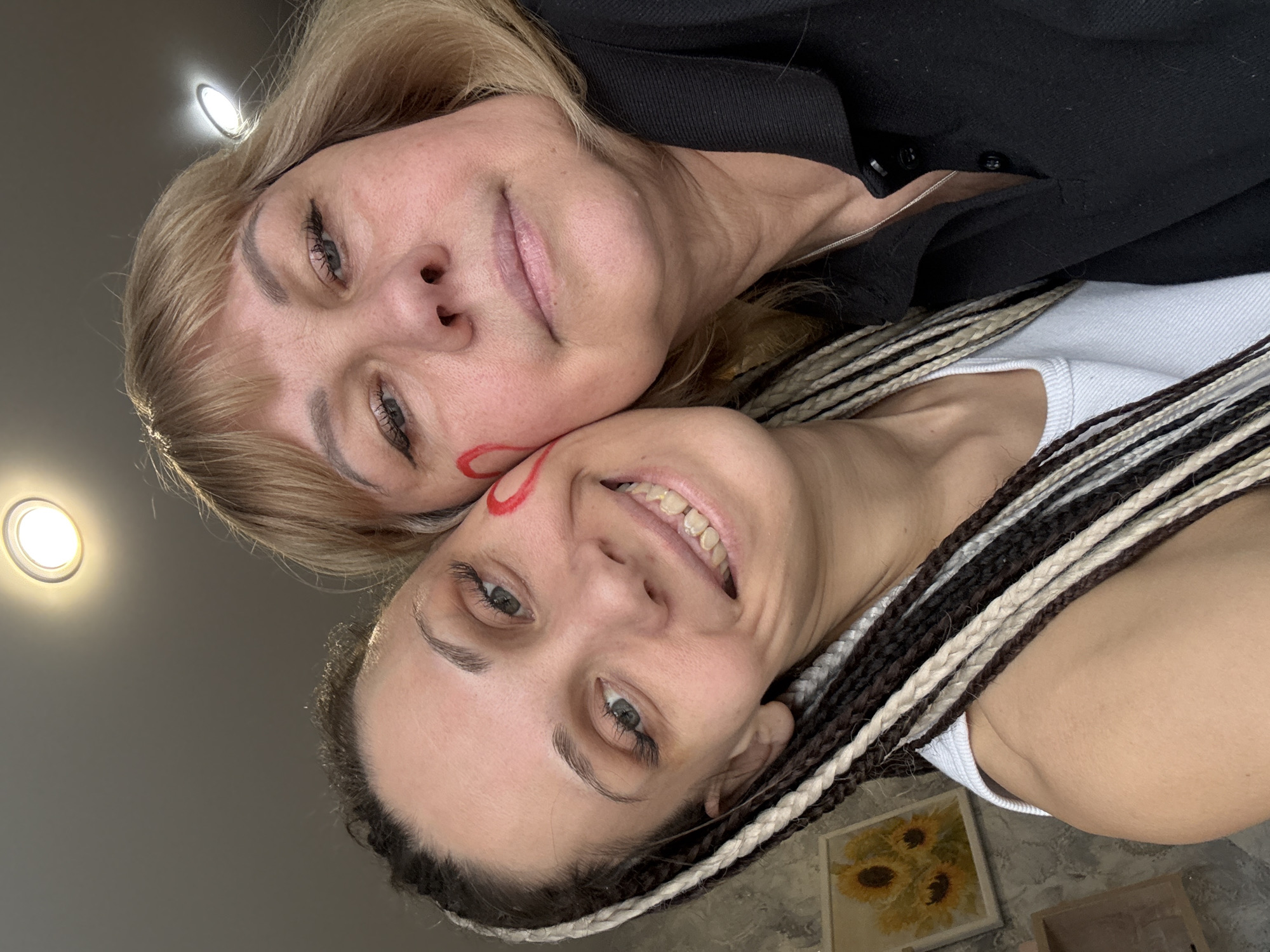 Two women are captured in a close-up, upward-angle portrait, likely a selfie. Their heads are resting together, occupying most of the frame. The woman on top, appearing older, has blonde hair styled with bangs and is wearing a black collared shirt. She has dark eyeliner and her lips are slightly parted in a gentle smile, looking directly at the camera. Her face is in soft focus.
Below her, a younger woman with dark and light braided hair (dreadlocks or box braids) smiles broadly, showing her upper teeth. She wears a white top and has a distinctive red, swirling mark or design on her left cheek. Her eyes are open and looking towards the camera. The women's faces convey a sense of warmth and connection.
The background is a plain, light grey wall or ceiling, with two bright, round recessed ceiling lights visible, casting a glow. In the lower right corner, behind the younger woman, a framed picture is visible on the wall, depicting what appears to be yellow sunflowers. The setting is indoors, possibly a home or a personal space in Rusanovo, Russia. There is no visible text in the image.