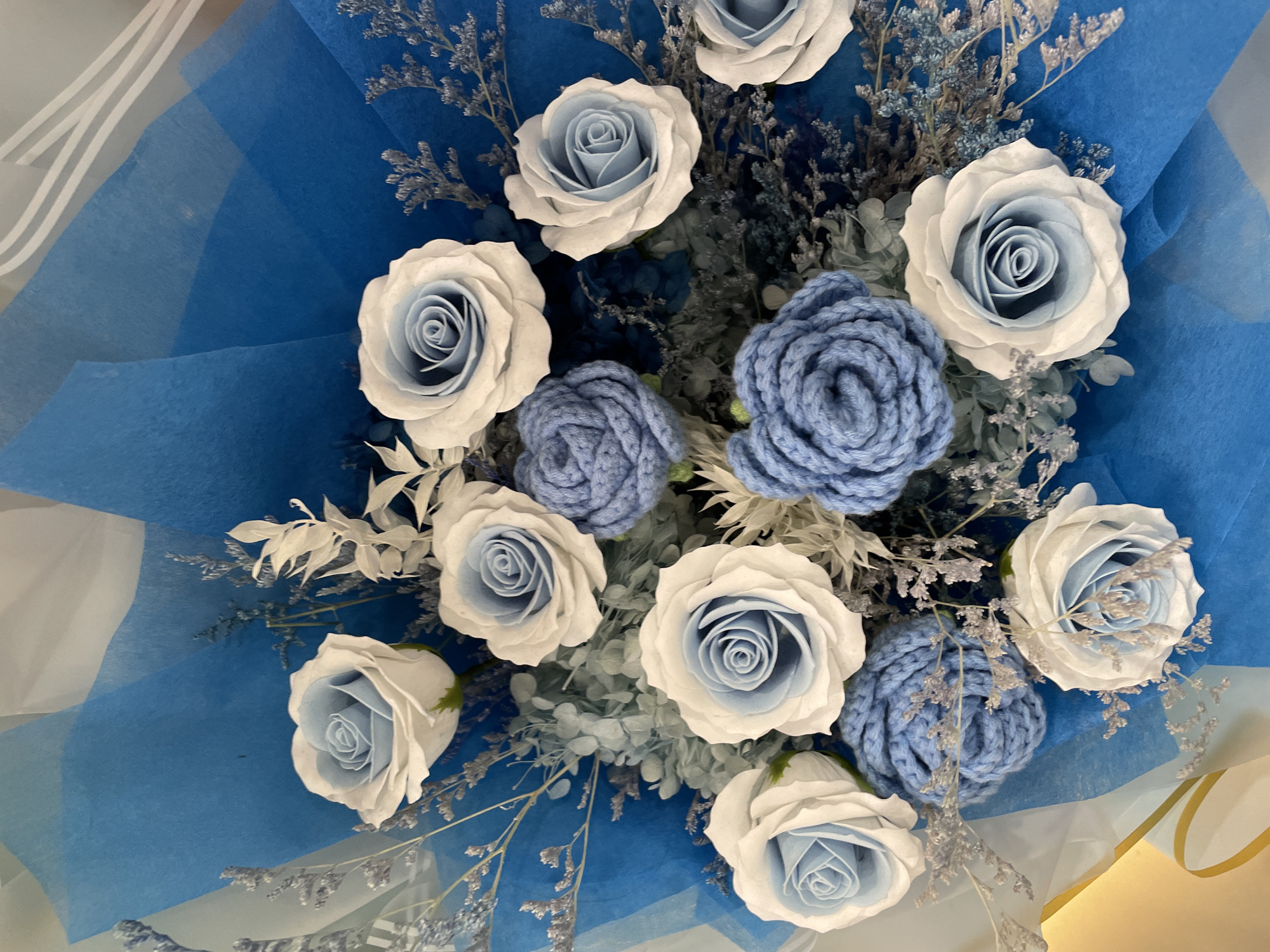 A close-up, overhead shot captures a meticulously arranged bouquet predominantly featuring blue and white tones. The central subjects are several roses; some have light blue centers fading to white outer petals, while others are distinctly crafted from blue yarn, resembling crocheted roses. These are interspersed with delicate, wispy filler elements in shades of silver-grey and muted blue-green, possibly dried or preserved flowers and foliage, adding texture and depth to the arrangement.
The bouquet is wrapped in multiple layers of paper, primarily a vibrant royal blue, with some translucent white or very light blue layers providing a soft contrast. The crinkled texture of the wrapping paper is visible, contributing to the overall aesthetic. Part of a gold or yellow surface, possibly a ribbon or another part of the wrapping/container, is visible at the bottom right corner of the frame.
The arrangement appears dense and full, filling the majority of the frame. The lighting is soft and even, highlighting the different textures of the petals, yarn, and dried elements without harsh shadows. No specific activity is depicted, as it is a still life of the bouquet, likely prepared for gifting or display. There is no visible text in the image.