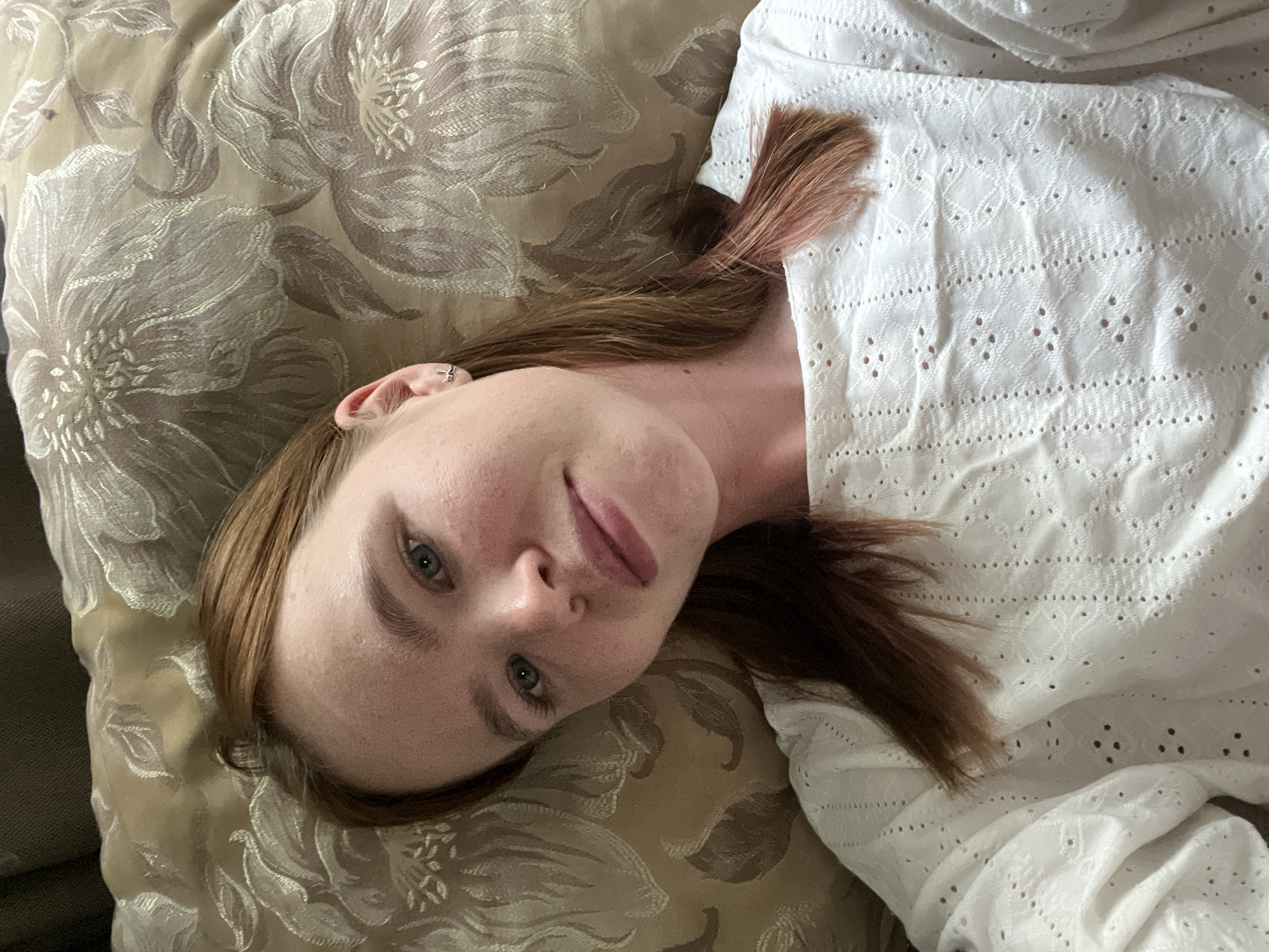 A young woman with auburn hair is lying on a pillow with a floral pattern. She is wearing a white, eyelet-style blouse. Her head is tilted towards the camera. The location is unspecified and cannot be identified from the image.