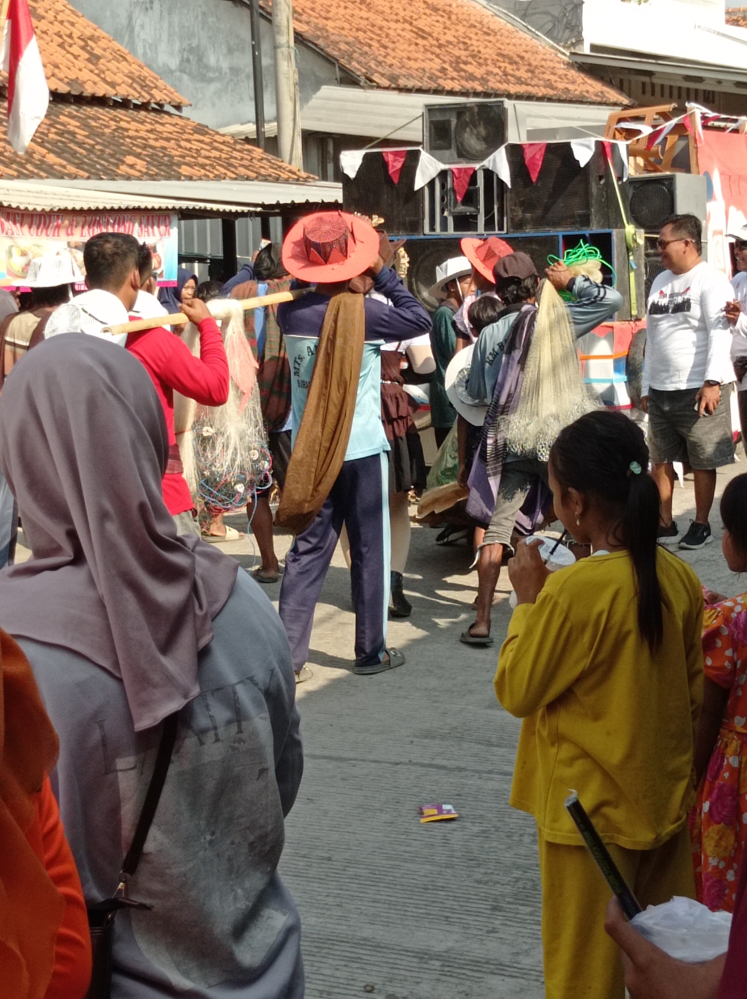 The image depicts a street scene in Maribaya, Indonesia, likely during a local event or festival. In the foreground, a person wearing a purple hijab and a grey shirt is visible from the back, with a black strap of a bag visible across their chest. To their right, a young girl in a bright yellow long-sleeved shirt and pants is holding a white cup and a dark object. Behind her, another person is partially visible wearing a patterned orange and pink dress.
In the mid-ground, several individuals are seen. A person in a red shirt carries a long wooden pole across their shoulders, from which a fishing net with various items attached hangs. Another person is wearing a distinctive wide-brimmed red hat and a blue and dark blue outfit. Others are dressed in casual attire.
The background features buildings with red-tiled roofs, some adorned with Indonesian flags. A large sound system with multiple speakers is set up, decorated with red and white triangular flags. The ground is paved with concrete. The overall atmosphere suggests a lively outdoor gathering.