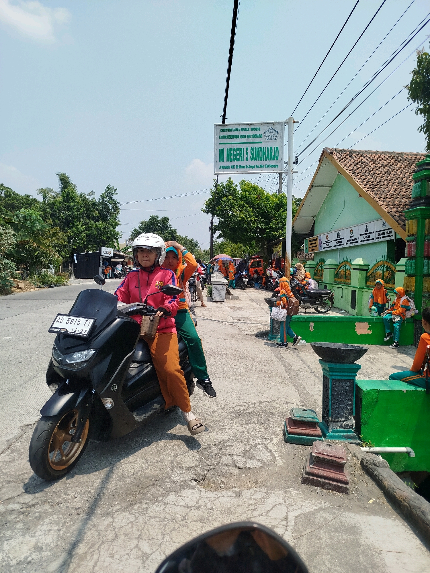 Two women on a black scooter are the main subjects in this image, taken on a sunny day. The woman in front is wearing a pink jacket and an orange headscarf, while the woman behind her has on an orange jacket and a green headscarf. They are both wearing casual clothing. The scooter has a license plate that reads "AD 5815 TT" and a time stamp of "12:27".
In the background, there is a building with a green facade and a tiled roof, identified by a sign as "MI NEGERI 5 SUKOHARJO" located on "Jl. Wonodadi Km.7 Ds.Grogol Kec. Miri Kab. Sukoharjo". Several children, presumably students, are seen near the building, some wearing orange and green uniforms. They are sitting on a low green wall and walking around. The environment appears to be a street with a sidewalk, trees, and power lines overhead. The overall scene suggests a school environment in the Sukoharjo regency of Indonesia, specifically in the Grogol sub-district.