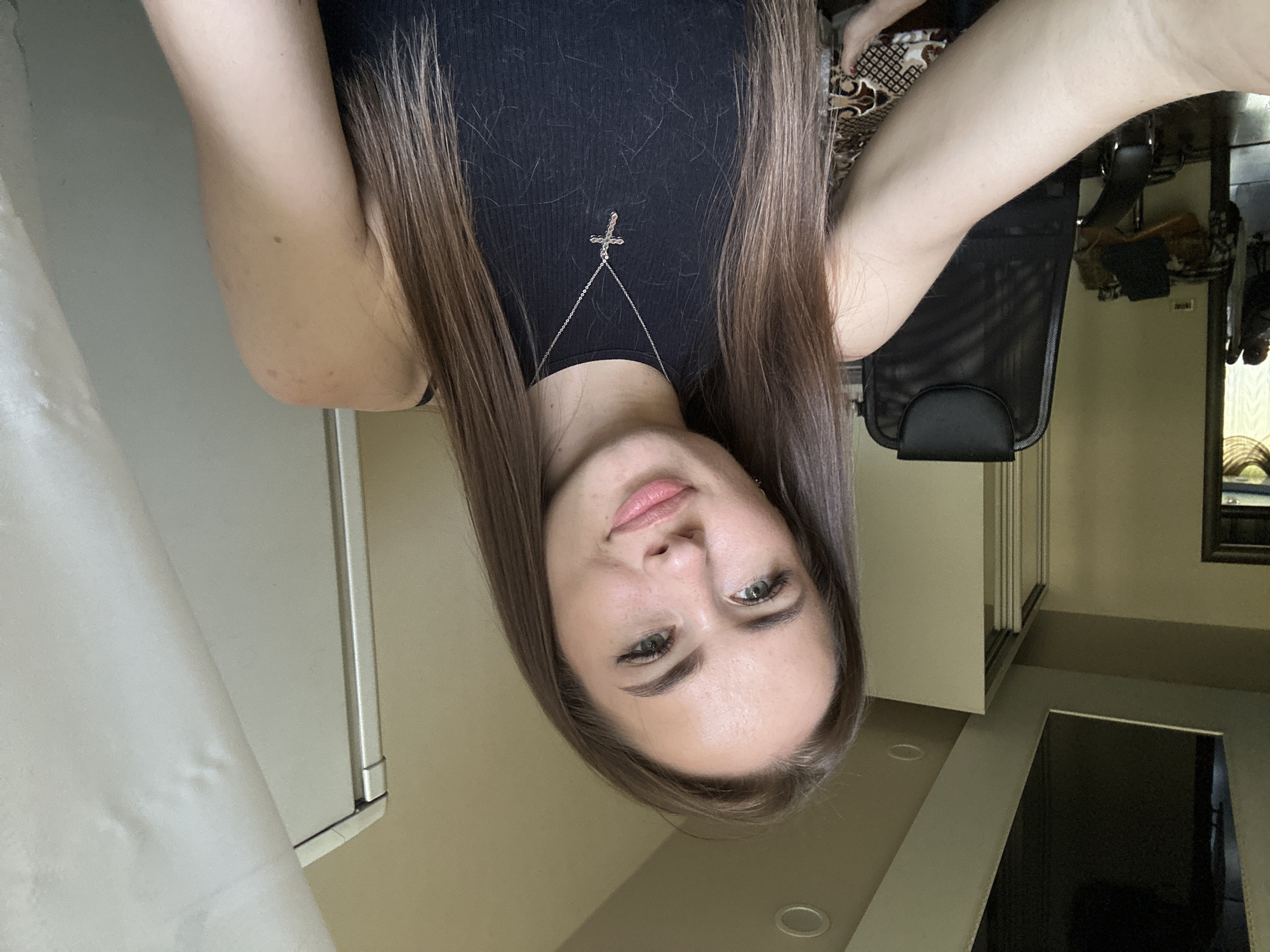 A female with long, straight, brown hair featuring lighter ends, consistent with an ombre or balayage style, is centered in a medium close-up selfie, looking directly at the camera with a slight smile. She has light skin, green eyes, and light eyebrows. She is wearing a black ribbed sleeveless top and a silver chain necklace with a silver cross pendant. Her right arm is partially visible on the left side of the frame, angled towards the camera.
The background depicts an indoor residential or office setting. To the subject's left, a light beige wall extends into the distance, revealing a dark wooden doorframe or archway. Beyond this archway, a dimly lit room is visible. Closer to the subject, a black mesh-backed office chair with a headrest is positioned. Partially obscured below and to the left of the chair is a surface covered with a patterned brown, white, and black geometric cloth. The ceiling above the subject's left side is light beige and features a large, dark rectangular recessed area, possibly a skylight or a reflective panel, and a circular recessed light fixture.
To the subject's right, a light beige wall transitions into a vertical, light-colored structure, possibly a cabinet or a part of a wardrobe, which has a prominent silver vertical trim on its left edge. A white curtain or piece of fabric hangs partially in front of this structure. The ceiling above the subject's right side is also light beige and contains a circular recessed light fixture.
