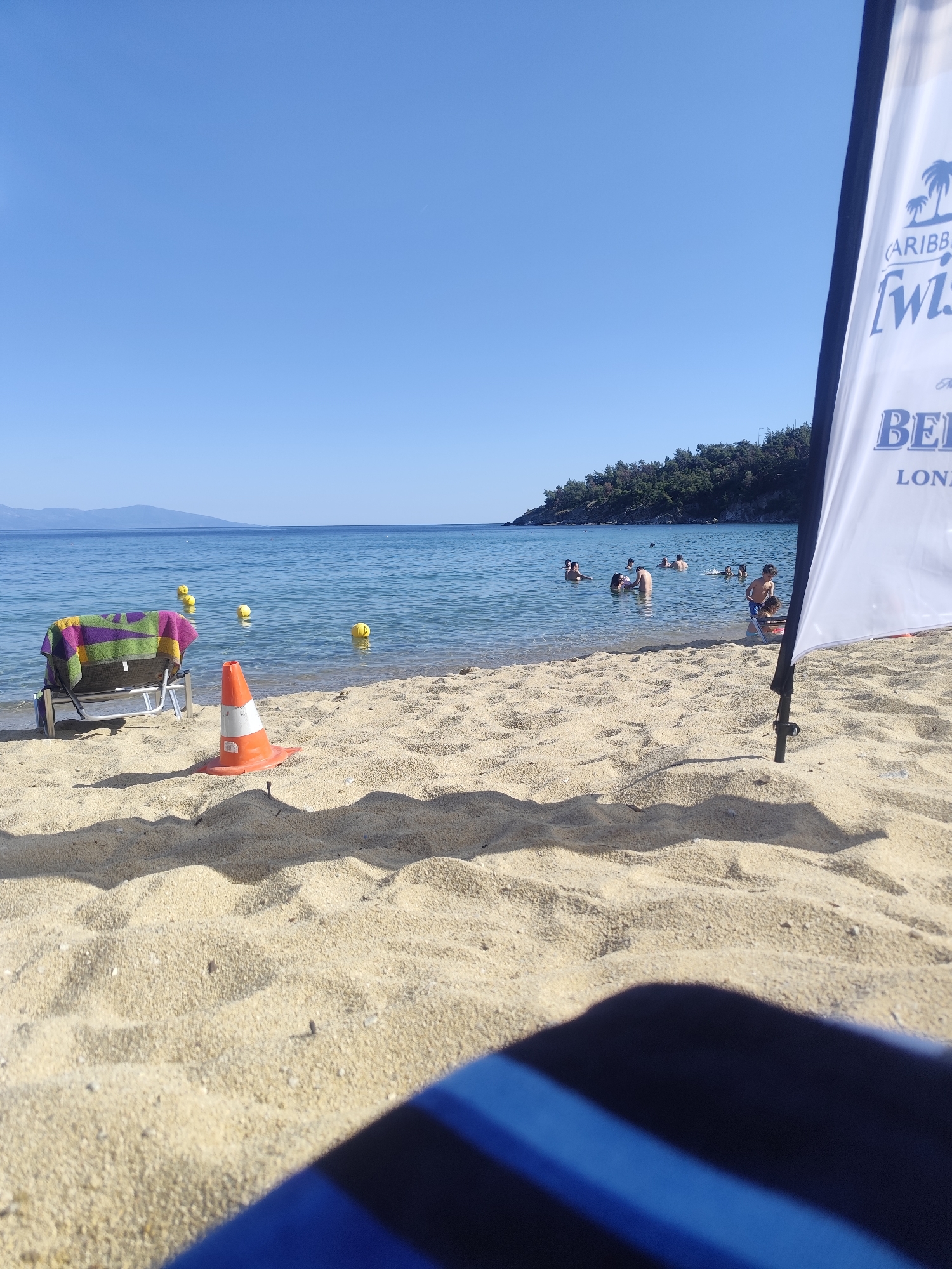 The image captures a sunny day at a beach, likely in or near Kavala, Greece. The foreground is dominated by sand, with the blurred blue and black stripes of what appears to be a beach towel or chair in the bottom right corner. A bright orange and white traffic cone stands prominently on the sand in the mid-ground, suggesting some sort of demarcation or temporary measure. To the left, a folded beach chair is draped with a colorful striped towel.
The water is calm and clear, with several yellow buoys floating on the surface. In the distance, a small group of people are visible swimming and wading in the sea. The shoreline is backed by a lush green hillside covered in trees, with mountains visible in the far distance under a clear blue sky. A white flag with blue text and imagery, possibly a logo or advertisement for "CARIBBEAN TWIST" and "BELL", is partially visible on the right side of the frame. The overall scene depicts a relaxed beach atmosphere on a warm day.