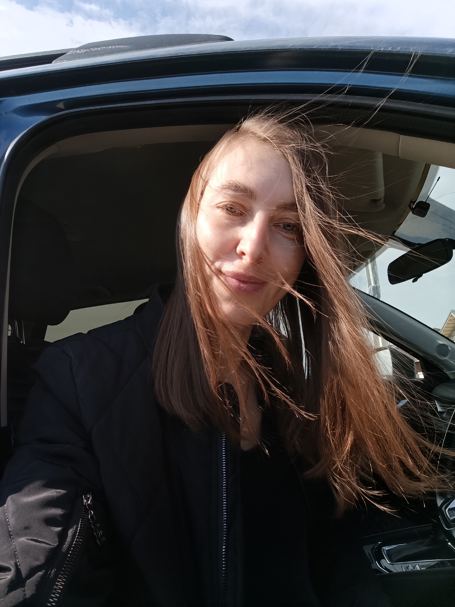 A selfie shows a woman with long, straight, light brown hair, some strands of which are blowing across her face. She has fair skin and a neutral expression. She is wearing a black jacket and appears to be sitting inside a dark-colored vehicle, possibly a car, with the door open. The interior of the vehicle is visible, including the steering wheel, dashboard, and rearview mirror. The sky is visible through the car window and above the car, showing patches of light blue with some white clouds. The reflection in the car window shows a building exterior with a roofline, suggesting an urban environment. Given the request for a location in Chaikovsky, Russia, it can be assumed that the photo was taken in or near that city.