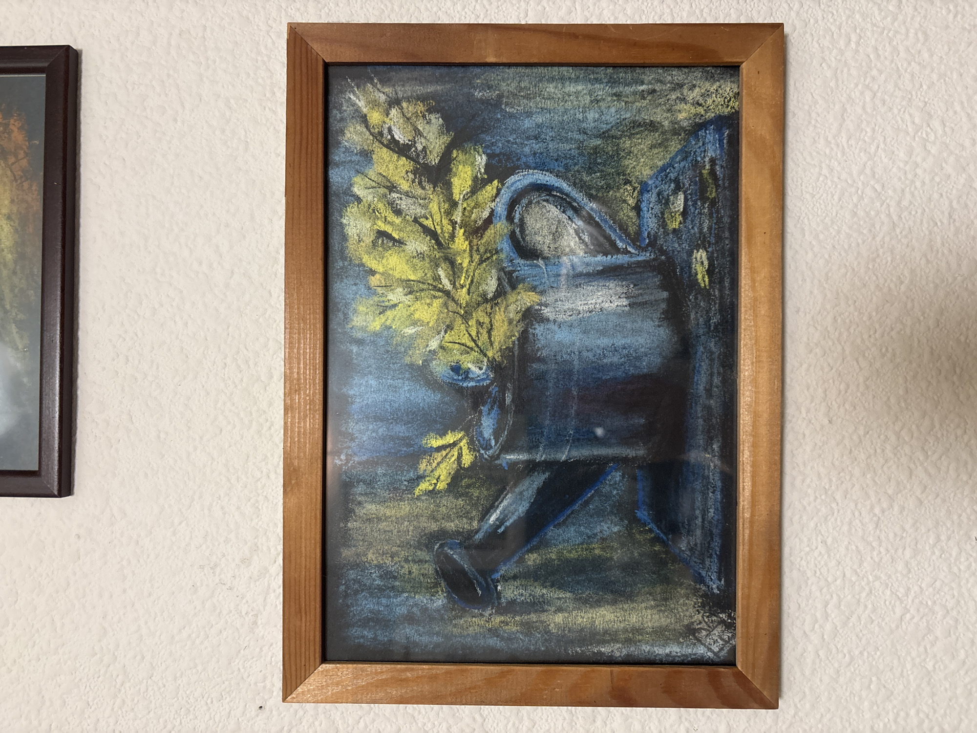 A framed pastel drawing of a blue watering can with yellow foliage is displayed on a textured white wall. The watering can is on a dark blue surface, possibly a table or shelf. The background is a mix of dark blue, green, and yellow, suggesting a textured or abstract environment. The drawing is in landscape orientation and has a thin wooden frame. Above and to the right of the main artwork, a portion of another framed picture with a dark frame is visible against the white wall. The image is taken from a slightly elevated angle, looking down at the artwork. There are no people or animals present in the image. The overall scene is still and appears to be a domestic interior.