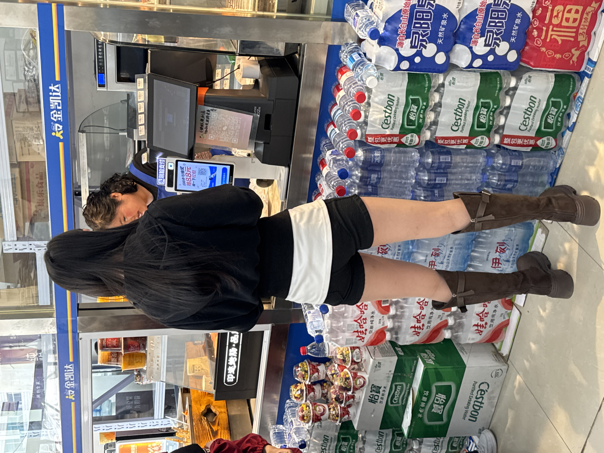 The image shows a woman standing at the checkout counter of a convenience store in Zhongtun, China. She has long black hair and is wearing a black top, white shorts, and brown boots. The checkout counter is stocked with various items, including bottled water (brands like Cestbon and Wahaha), yogurt, and packaged foods. The store's logo, "金凯达," is visible on the side of the counter. A digital payment screen with QR codes is in view. The environment appears to be a typical neighborhood convenience store.