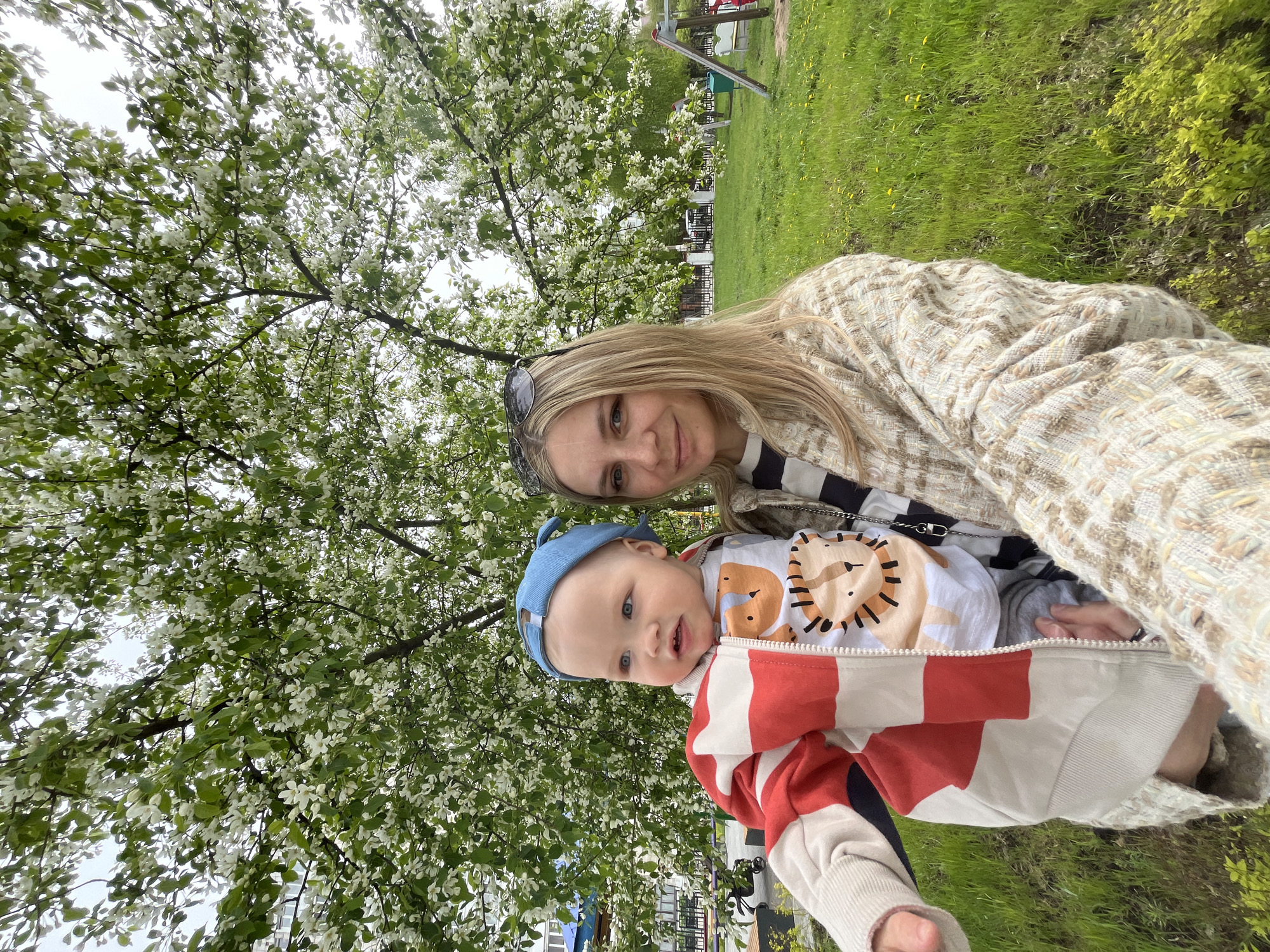 A woman and a baby are in an outdoor setting, possibly a park or recreational area in Chaikovsky, Russia, as suggested by the location. The woman has blonde hair and is wearing a jacket, while the baby is wearing a blue cap and a jacket with a red and white pattern. There is a tree with white blossoms in the background. The environment includes green grass and playground equipment in the distance. The image appears to be a selfie.