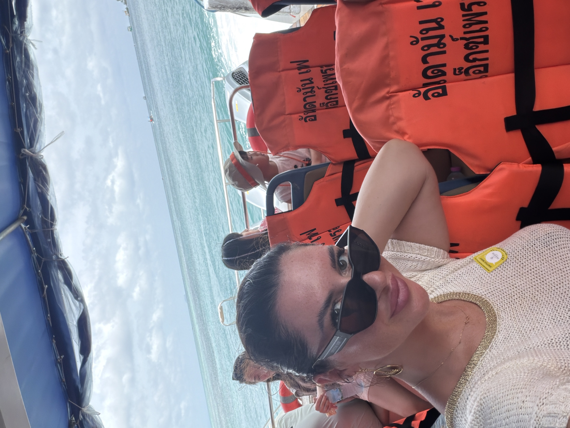 A woman with dark hair and sunglasses takes a selfie on a boat with other passengers. She is wearing a white knitted top with a gold trim. The boat has orange life vests on it. The water appears to be the sea, with other boats in the distance under a cloudy sky. The location is Ao Nang, Thailand.