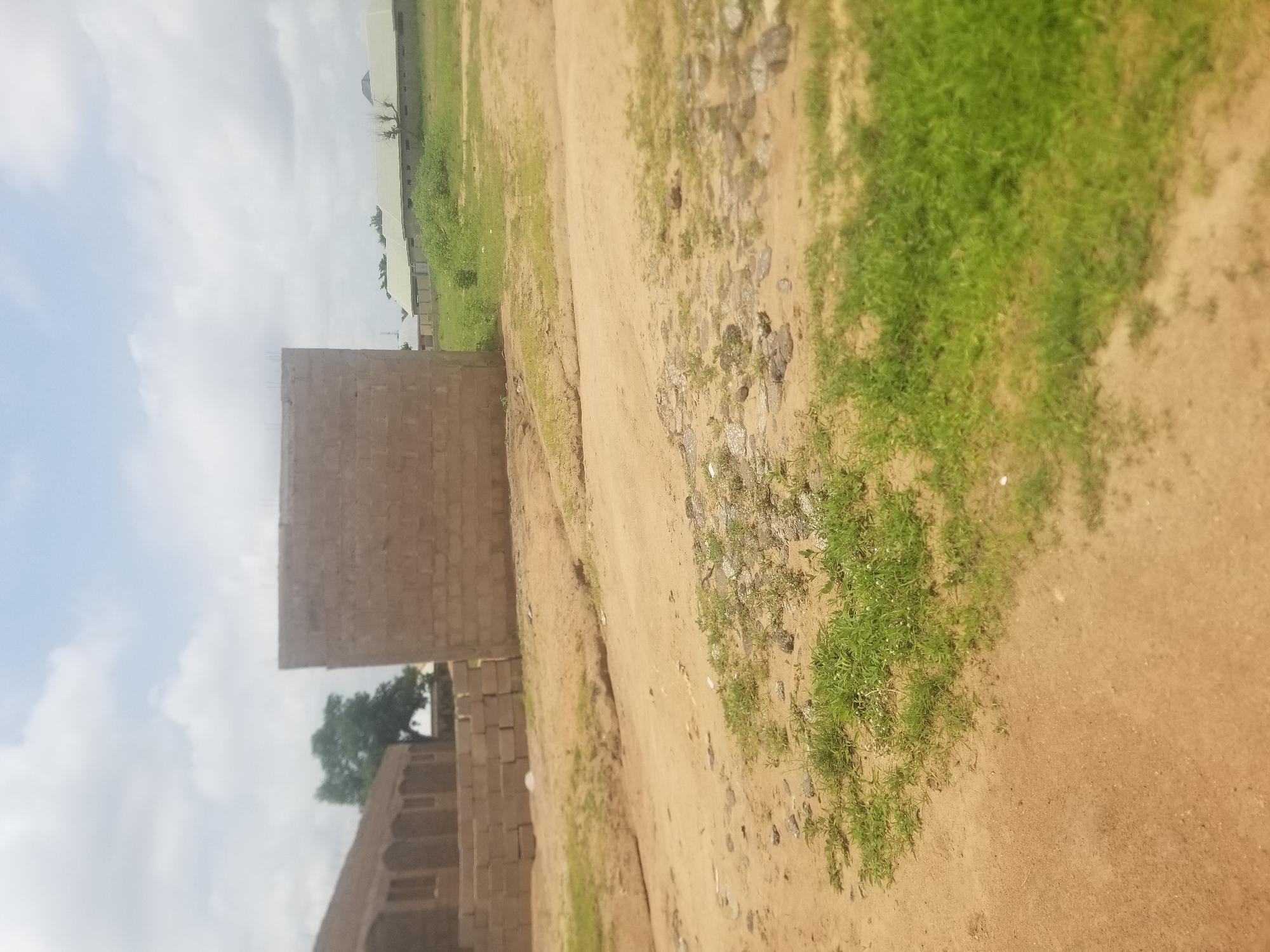 The image depicts an outdoor scene in Jauro Yinu, Nigeria, characterized by a dirt road with sparse green vegetation and scattered rocks. In the background, several structures are visible. On the left, a building with arched windows and a brick facade is under construction. To its right, a large, unfinished concrete block structure stands prominently. Further in the distance, a row of buildings with light-colored, sloped roofs stretches across the landscape. The sky above is overcast with white clouds.