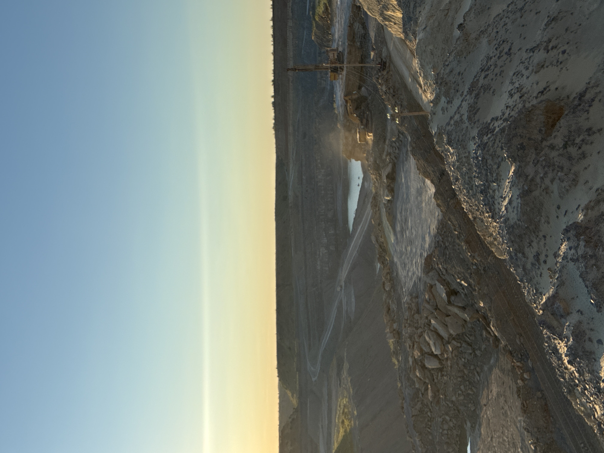 This media file captures a wide, panoramic view of an open-pit mine, likely a quarry, in Tatsinskaya, Russia. The scene is dominated by the layered rock formations and excavated earth, indicative of large-scale mining operations. In the mid-ground, a yellow mining vehicle, possibly a drill rig or loader, is visible. A body of water, likely collected rainwater or a pond, is situated at the bottom of the quarry. The overall impression is one of industrial activity against a natural, rugged landscape.
The time of day appears to be late afternoon or early evening, given the warm, golden light illuminating the upper sections of the quarry and casting long shadows. The sky transitions from a clear, pale blue on the left to a hazy, yellowish hue towards the horizon on the right, suggesting a sunset or sunrise. The weather appears clear, with no visible precipitation or overcast conditions.
Notable details include the visible stratification of the rock walls, suggesting different geological layers that have been extracted. Winding roads or tracks are visible throughout the quarry, indicating the movement of heavy machinery. The texture of the exposed rock surfaces is rough and varied, with some areas showing sharp, angular fragments and others appearing more weathered and eroded. The image conveys a sense of scale and the immense effort involved in extracting resources from the earth. No visible text is present in the media file.