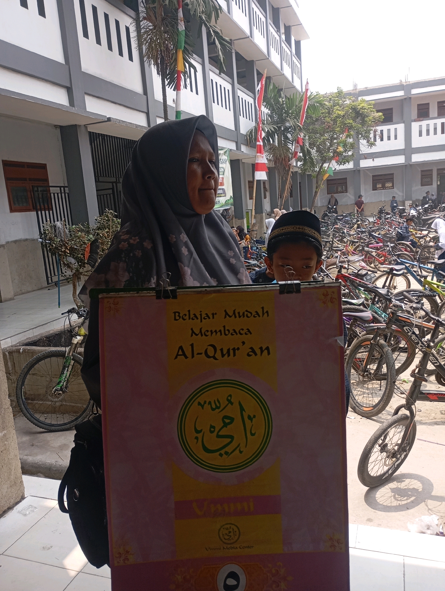 In Babelan, Indonesia, a woman wearing a grey hijab is holding a large sign with Arabic calligraphy and text in Indonesian. The sign appears to be educational, with the words "Belajar Mudah Membaca Al-Qur'an" which translates to "Easy to learn to read the Quran." Beside her, a young boy wearing a black cap with gold embroidery peeks over the sign. The background reveals a school building with a white and gray facade, several palm trees with Indonesian flags, and a large number of parked bicycles. The overall scene suggests an outdoor event or gathering, possibly at a school or community center, with bicycles indicating a mode of transportation for many attendees. The ground is paved with tiles, and there is a concrete gutter with a bicycle parked next to it. The lighting suggests it is daytime.