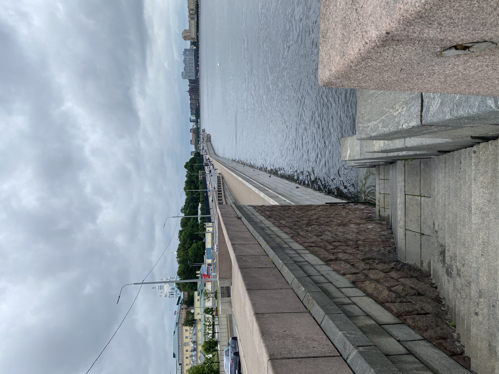 The image shows a wide, grey river on a cloudy day, with a stone embankment along its left side. A set of stone stairs descends from the embankment into the water. On the embankment, a paved walkway is visible, with a granite wall separating it from the river. Beyond the walkway, a road with cars and streetlights runs parallel to the river. In the background, several buildings are visible, including a white church with blue domes and a row of multi-story residential buildings. The sky is overcast with dark grey clouds.