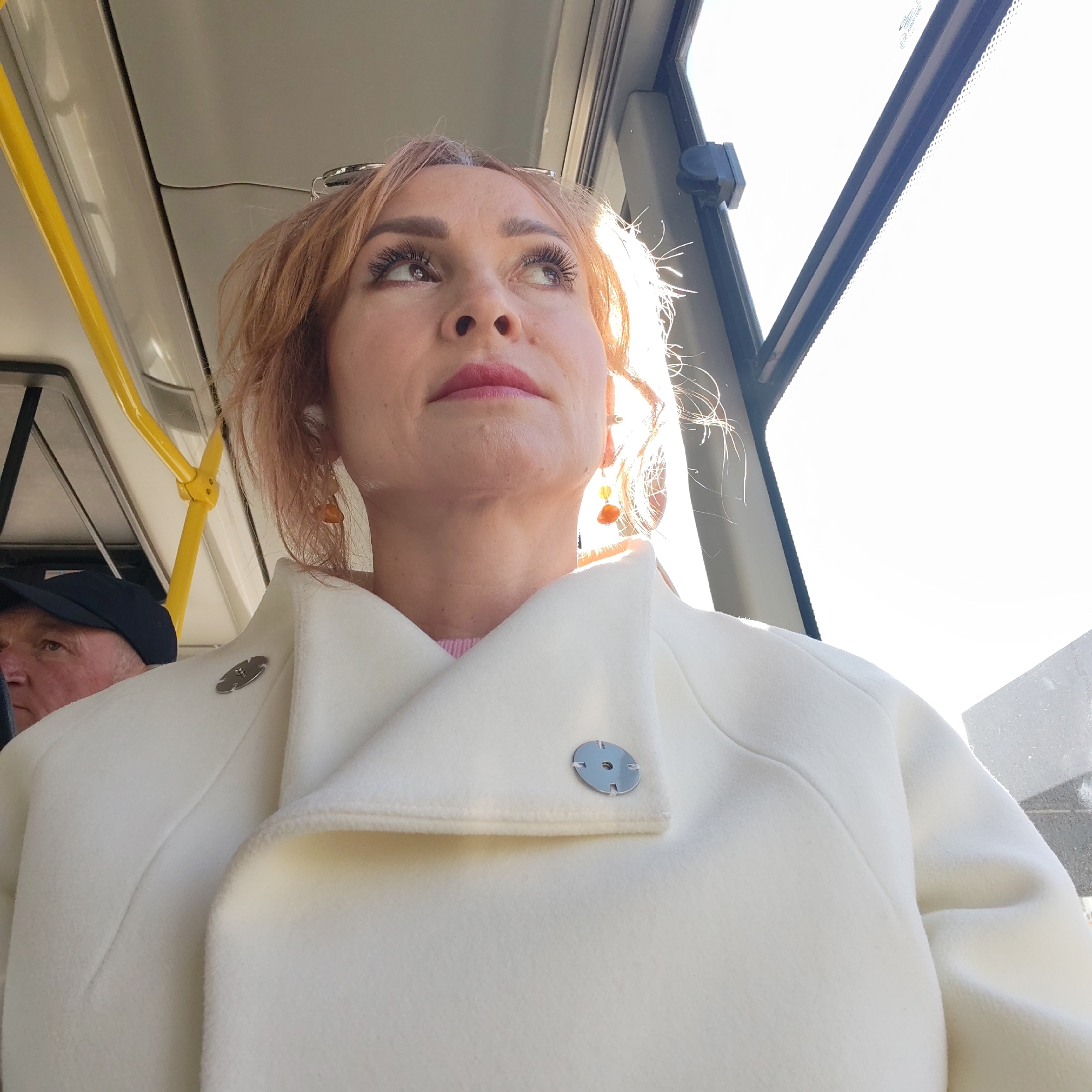 A fair-skinned woman with reddish-blonde hair, wearing a white coat with round metallic buttons, sits on a bus in Chaikovsky, Russia. She is looking up and to the right. An older man wearing a black cap sits to the left. A yellow handrail is visible in the background. Sunlight streams through the bus window to the right. The scene appears to be inside a public transport vehicle in Chaikovsky, Russia. The woman has orange earrings and appears to be wearing wireless earbuds.