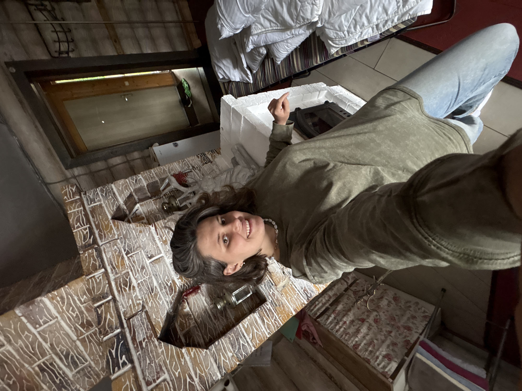A woman is lying on her back on a tiled floor, facing the camera. She is wearing a green sweatshirt and blue jeans. Her hair is dark brown and appears wet. She is smiling and pointing her right index finger upwards. In front of her is a white, styrofoam-like object with a black metal insert, possibly a fireplace surround. Behind her, a bed with white bedding is visible. The setting appears to be an attic or a room with a sloped ceiling and a skylight. The walls are made of brick or stone with a white pattern. The image has a slightly grainy texture. The lighting suggests it is daytime, possibly indoors with natural light from the skylight. The overall mood seems relaxed and candid. No visible text is present in the image.