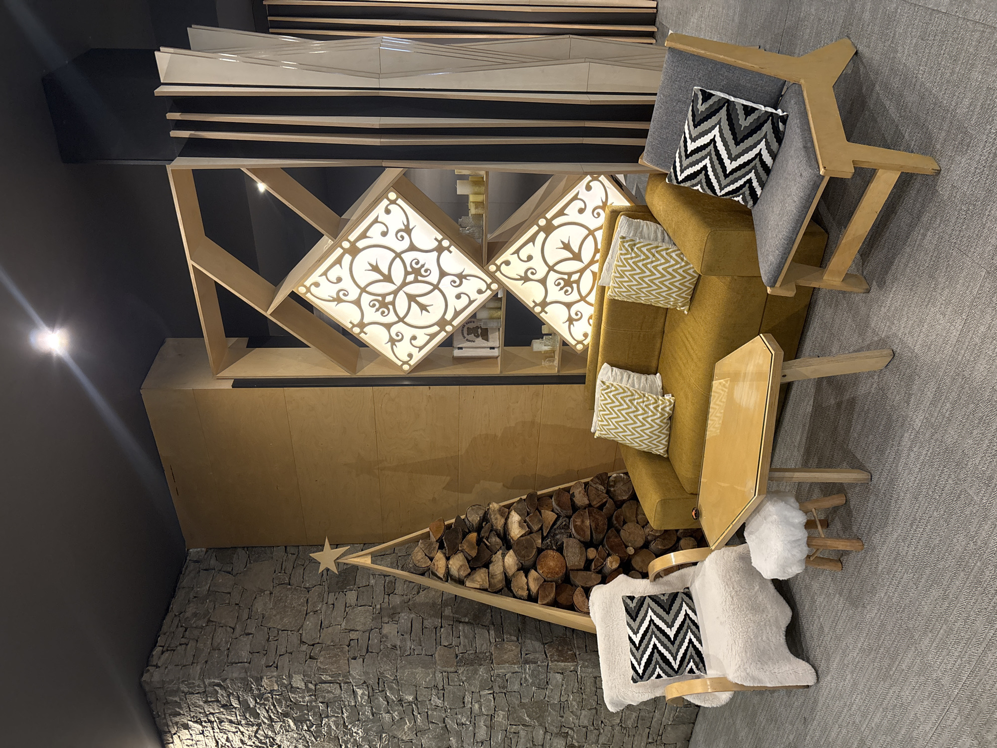 This image captures an indoor setting, possibly a lounge or waiting area. The floor is covered with a neutral-colored carpet. A feature wall is constructed from rough stones, and a structure resembling a Christmas tree made from wood and filled with logs is placed in front of it. Above the tree is a shelf or counter, over which are two decorative diamond-shaped lighting panels with intricate patterns.
To the right of the frame is an upholstered armchair with wooden framing and legs, accompanied by decorative pillows. A smaller wooden stool or side table with a plush cushion is nearby. On the wall above the lighting panels, there's a design feature of stacked materials. The overall environment suggests a modern, design-focused interior, with natural elements and unique lighting.