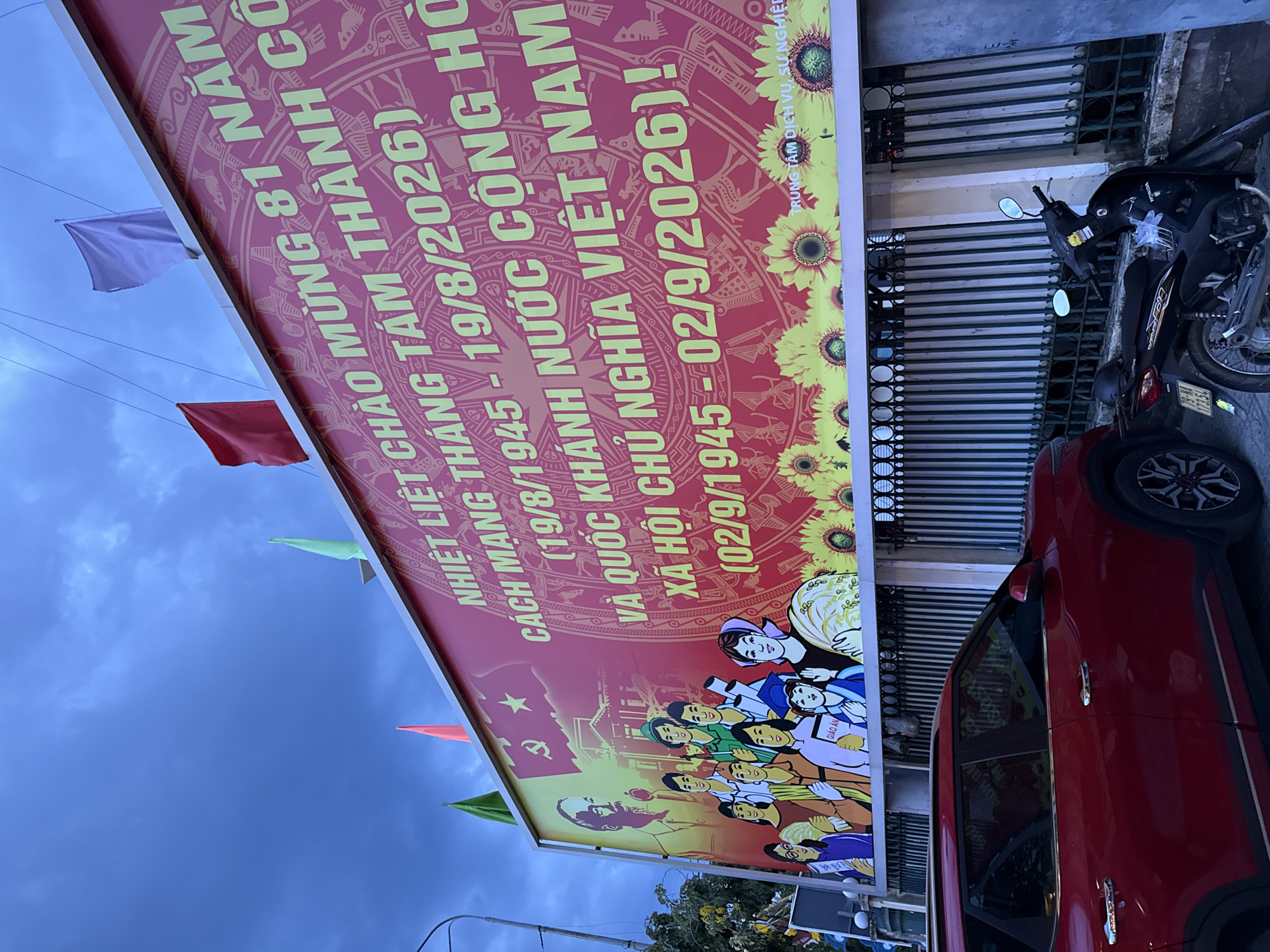 The image shows a large red banner displayed on the exterior of a building, likely in Nha Trang, Vietnam, given the Vietnamese text. The banner is adorned with yellow lettering and imagery, celebrating the 81st anniversary of "CÁCH MẠNG THÁNG TÁM" (August Revolution) and "QUỐC KHÁNH NƯỚC CỘNG HÒA XÃ HỘI CHỦ NGHĨA VIỆT NAM" (National Day of the Socialist Republic of Vietnam). Specific dates mentioned are August 19, 1945, and September 2, 1945, with a future date of 2026 indicated, suggesting an upcoming celebration or memorial. The banner features a stylized illustration of a group of people, possibly representing Vietnamese citizens, and a prominent image of a historical figure, likely Ho Chi Minh. A red star with a hammer and sickle emblem is also visible, signifying the communist ideology. The banner is decorated with a border of sunflowers at the bottom.
In front of the building, a red SUV is parked, and a black motorcycle is positioned next to it. The scene appears to be outdoors, with a cloudy sky overhead. The building itself has a metal fence with vertical bars in front of it. Electrical wires are visible above the banner. The overall impression is a public display commemorating a significant national event.