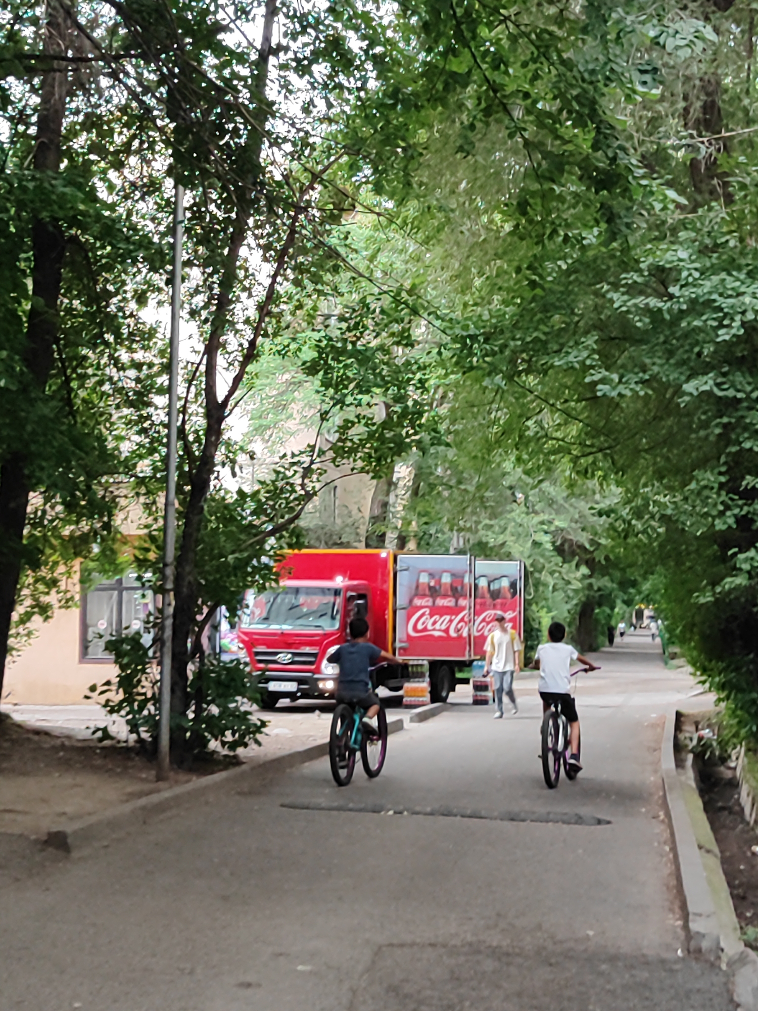In Almaty, Kazakhstan, a red Coca-Cola delivery truck is parked on a paved path lined with trees. Two boys are riding bicycles on the path. The boy in the foreground is on a turquoise and purple bike, facing away from the camera. The other boy, further ahead and to the right, is on a black and white bike, also facing away. A woman in a white shirt and jeans is walking in the background, near the truck. The scene appears to be during the daytime, with the trees casting shadows. The overall atmosphere is casual and outdoorsy. The Coca-Cola logo is clearly visible on the side of the truck, along with images of Coca-Cola bottles.