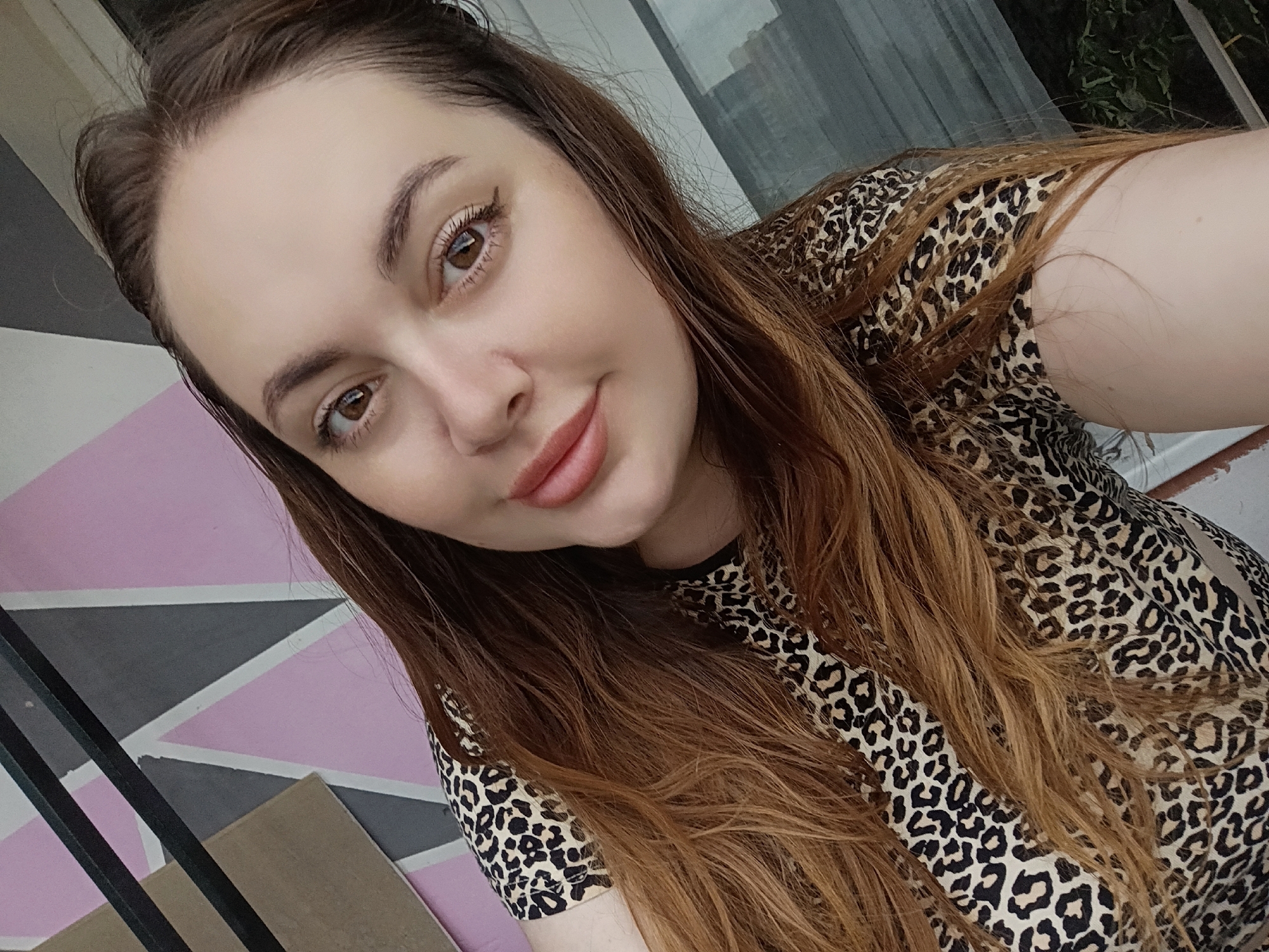A woman with long, light brown hair is taking a selfie. She is wearing a leopard-print top. Her eyes are brown and she is smiling slightly. The background is out of focus and appears to be a wall with a geometric pattern of pink, gray, and white triangles, and some dark vertical bars. There is also a blurred view of greenery and a window in the background. The image has a slightly tilted perspective. The location is not identifiable within the city of Chaikovsky, Russia, as no landmarks or context clues are present in the image.