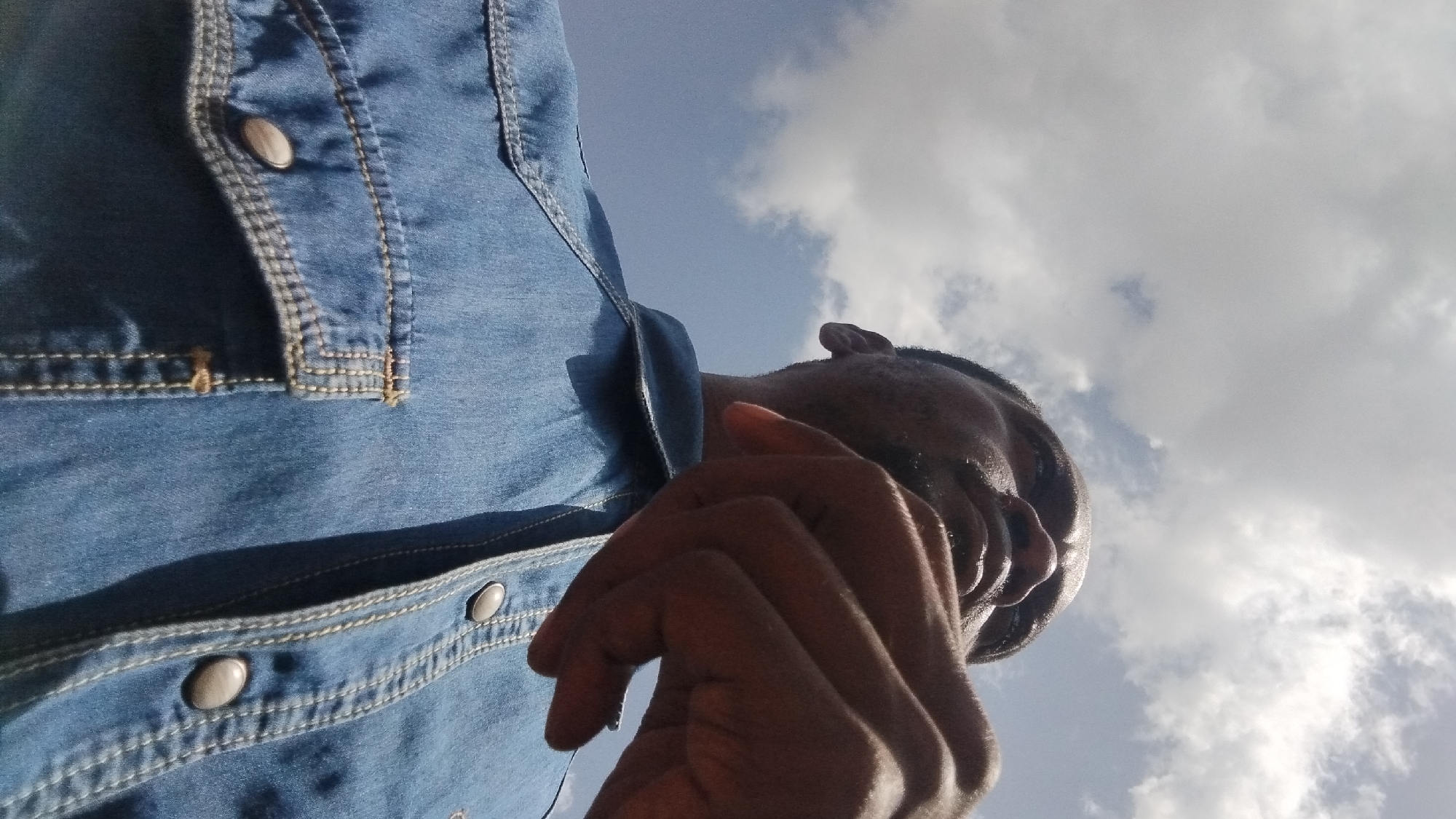 An upward angle shot shows a Black man wearing a blue denim shirt under a cloudy sky in Kabong, Nigeria. The man's face is partly obscured by his hand, which is held near his chin, and he is looking upwards. The denim shirt features prominent stitching and two buttoned chest pockets. The sky is a mix of blue and white clouds, suggesting an outdoor setting. The man appears to be outdoors, possibly in a public space within Kabong, Nigeria, given the context of the request.