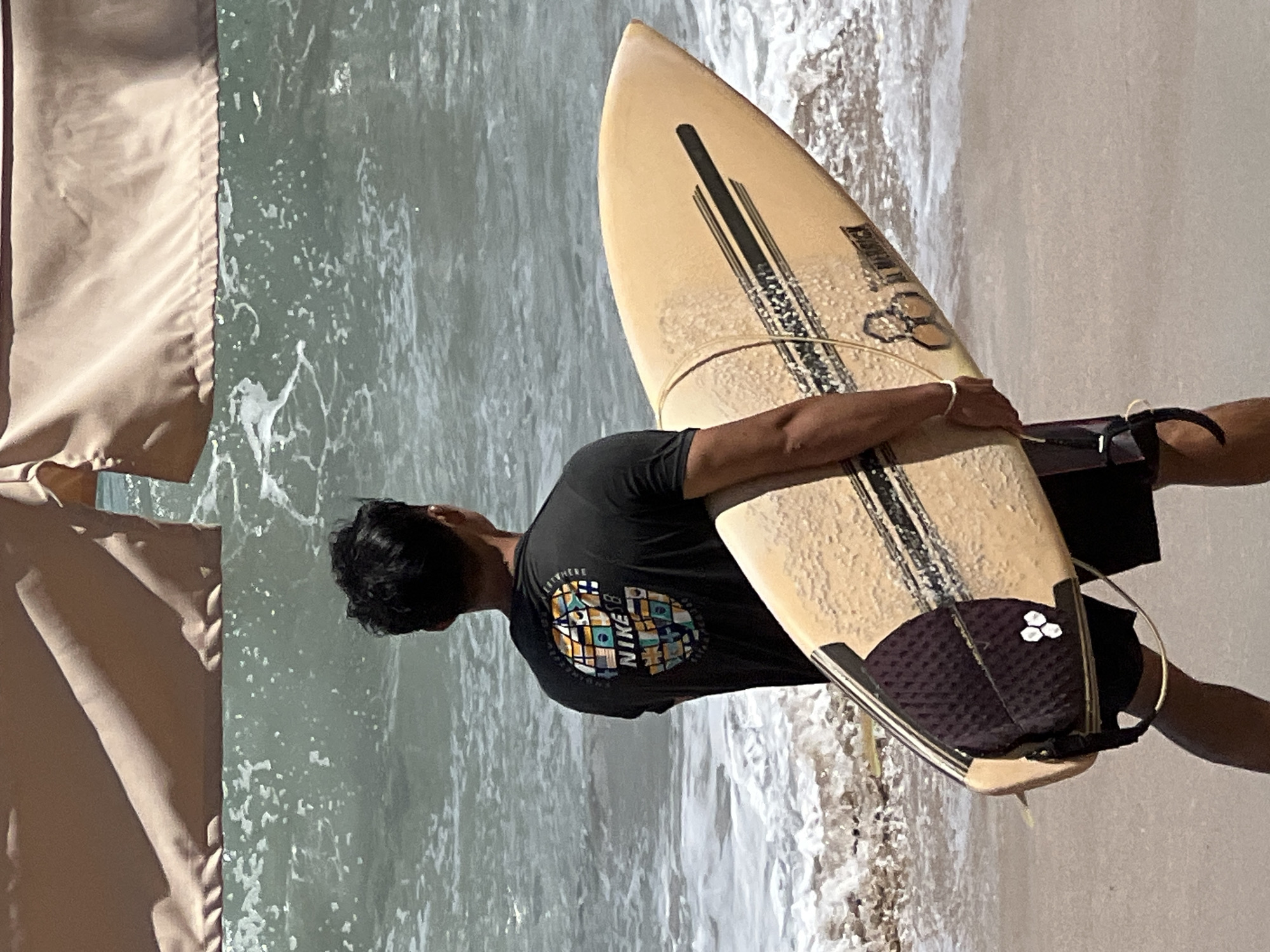 A surfer stands on the sandy shore of Kuta Beach, Indonesia, facing the ocean. He is holding a light brown surfboard with black accents and a purple traction pad. The surfer is wearing a dark t-shirt with a colorful graphic of a globe and the text "NIKE SB". The ocean behind him is choppy with white foam and breaking waves. In the background, part of a tan beach umbrella is visible.