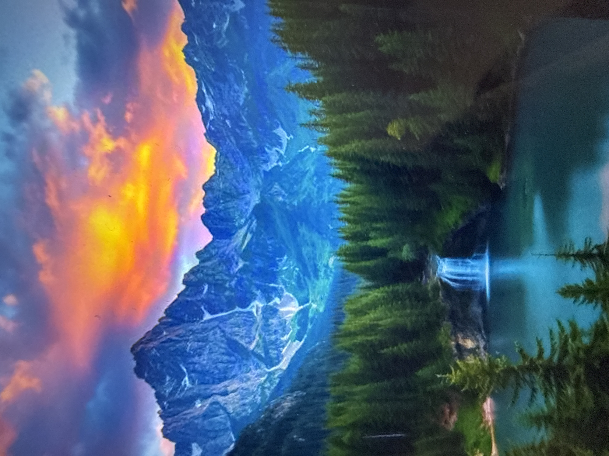This is a vibrant, high-angle, eye-level photograph of a majestic mountain landscape at sunset. In the foreground, a dense forest of dark green pine trees lines the edge of a tranquil, turquoise lake. A small, cascading waterfall flows into the lake from the right side of the frame. The middle ground is dominated by the lush green of the forest, with the lake reflecting the vibrant colors of the sky. In the background, imposing, jagged mountains rise steeply, their peaks dusted with white snow. The sky above is a dramatic spectacle of color, with fiery oranges and yellows blending into shades of pink, purple, and deep blue, suggesting a breathtaking sunset. The overall impression is one of serene natural beauty and dramatic skyscapes.