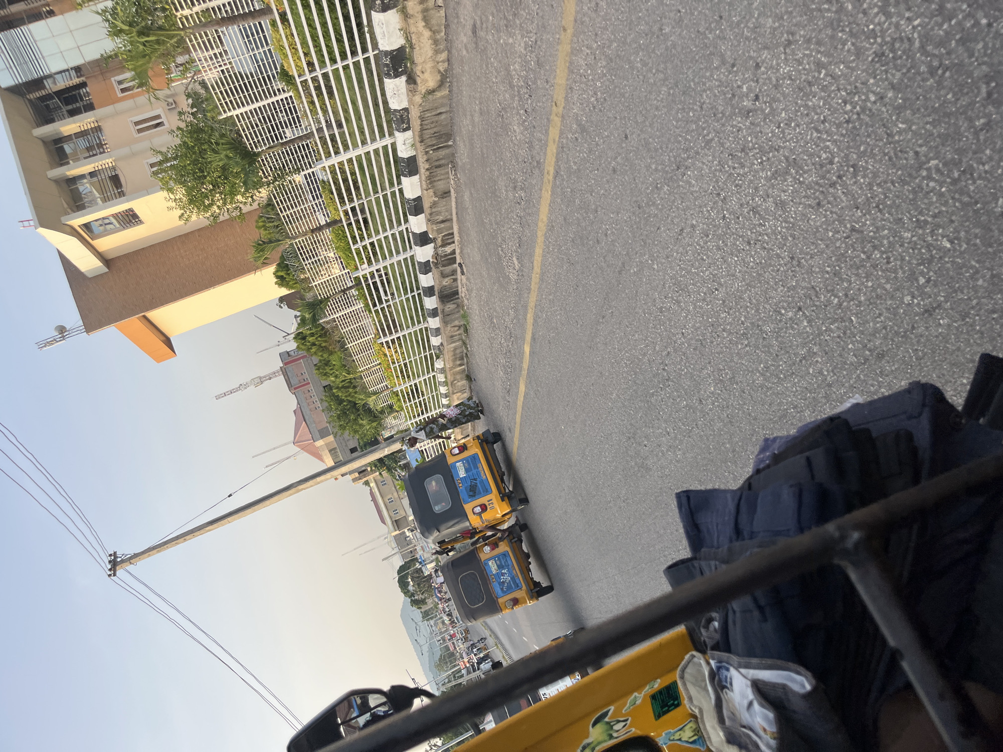 A low-angle, medium shot shows a street scene in a city, likely during daytime with a bright sky. In the foreground, a yellow auto-rickshaw's handlebars and some cargo are visible. Two other auto-rickshaws, also yellow with black tops, are in the middle ground, parked side-by-side on the right side of the road. The auto-rickshaws have visible license plates and some decals. To the right of the auto-rickshaws, a person is standing on the sidewalk, facing away from the camera and wearing a patterned dress. A modern, multi-story building with a glass facade and orange accents is located behind the auto-rickshaws, fenced by a white metal railing with black and white painted concrete barriers along the curb. A tall utility pole with wires is visible in the background to the left, along with other buildings, trees, and a distant mountain range. The road is asphalt with a yellow lane marker. The overall impression is a busy urban environment.