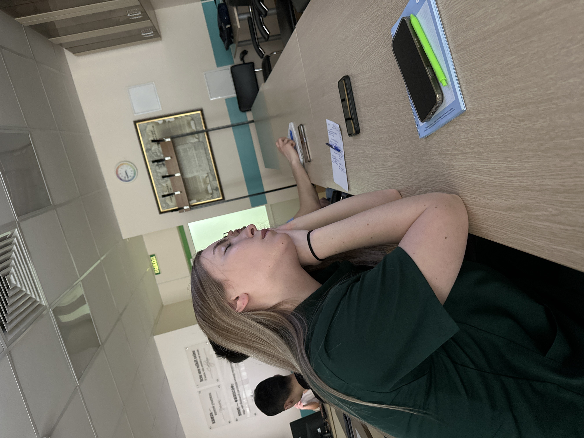 The image shows a woman with long blonde hair, wearing a dark green dress, resting her head on her arms on a light brown conference table. A man with short black hair is seated beside her, but partially obscured from view. Objects on the table include a black smartphone, a green stylus, a blue notebook, a black rectangular device, a pen, and a piece of paper with writing. The background includes a clock, a framed document or picture, and a row of coat hooks on a wall with a blue stripe. The wall is white with tiles. A sign with Cyrillic script and English spelling of the word “EXIT” is on the wall. The wall contains posted text that states “Kazan” and “Solo a mobil scito.”
The setting appears to be an office or conference room in Kazan, Russia.