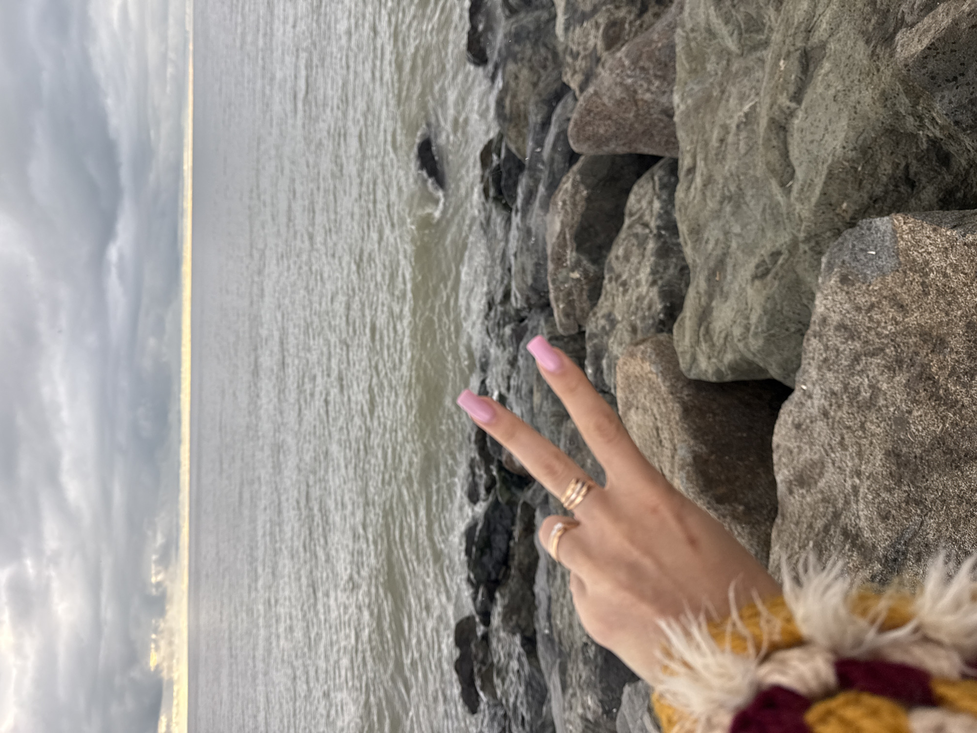 A vertical shot shows a woman's hand making a peace sign on a rocky shore, with the sea and sky in the background. The hand is light-skinned and adorned with rings, featuring pink-painted nails. The garment visible on the bottom right corner is patterned with yellow, white, and maroon colors and appears fluffy.
The background consists of gray, roughly textured rocks, leading to a gray sea with small waves. The sky is overcast with gray clouds. The picture appears to be taken outdoors. Although the location of Batumi, Georgia is mentioned, there is no way to confirm that based on the details shown in the picture.