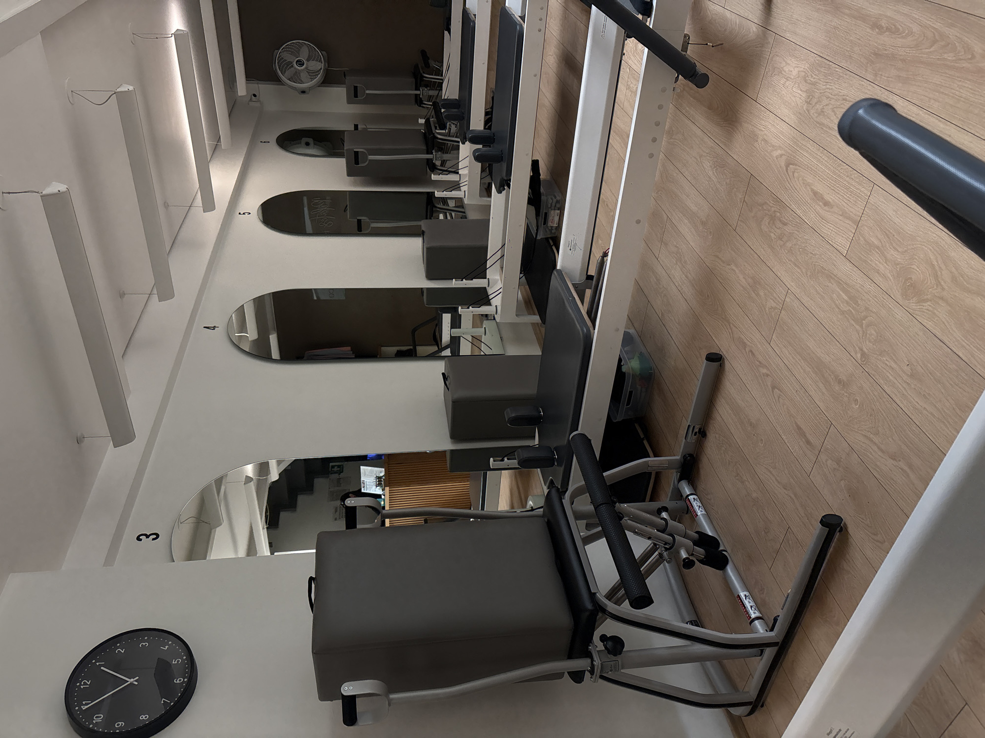 An interior shot depicts a modern and minimalist Pilates studio, likely located in Mexico City, Mexico. The room is equipped with multiple Pilates reformer machines, arranged in a row along a light wooden floor. White walls and vertical dividers create a clean, bright aesthetic, complemented by ample artificial lighting.
No people are present in the image, suggesting the studio is currently empty or between classes. The machines, primarily in shades of grey and white, are neatly aligned, ready for use. Each station is marked with a number on the white wall next to an arched mirror, with visible numbers including '3', '4', '5', and '6'. Reflections in the mirrors show more of the studio space, including another machine and what appears to be a staircase. On the mirror near station 5, faint reflections of text, possibly "LOVE&LIFE", are visible.
A black wall clock on the far left wall indicates the time is approximately 9:15. A small, gray desk fan is positioned on a shelf above station 6, suggesting temperature regulation. Small, possibly instructional or warning labels with text that appears to be in Japanese characters are faintly visible on the silver frames of some reformer machines. The overall atmosphere conveys a sense of calm and readiness for physical activity.