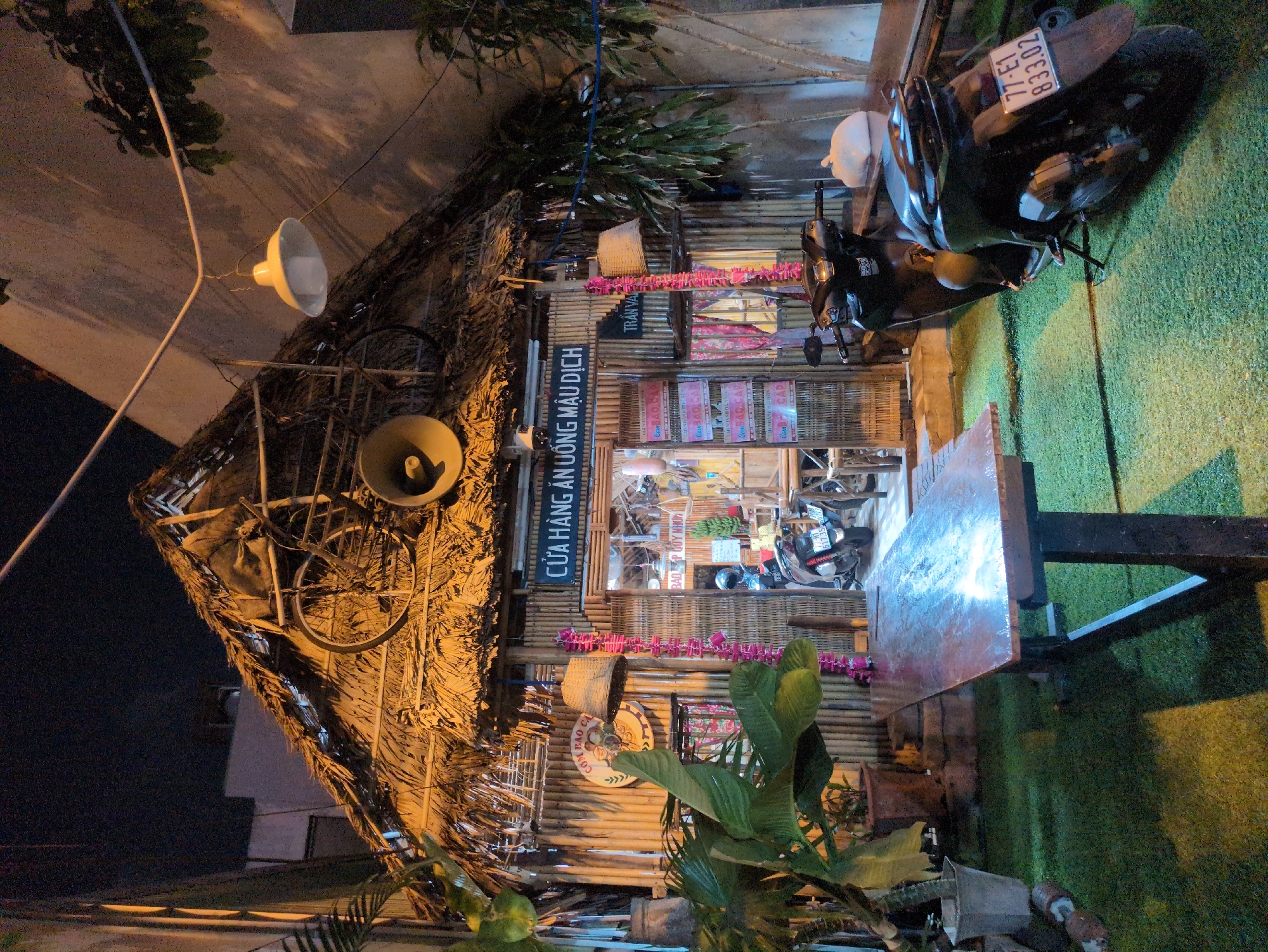 The image depicts a nighttime scene of a rustic, thatch-roofed establishment, likely an eatery or restaurant, located in South Quy Nhon Ward, Vietnam. The building features walls constructed from vertical bamboo poles and a prominent thatched roof. Positioned on the roof are a discarded bicycle frame and a large, light-colored horn-shaped speaker. Above the entrance, a sign in Vietnamese reads "CỬA HÀNG ĂN UỐNG MẬU DỊCH" (Food and Drink Mậu Dịch Store), with another sign displaying "CƠM BAO CÁ" and "BAO QUY NHON."
The immediate foreground features an artificial green turf flooring. A large, dark-stained wooden rectangular table is centrally located, with a white plastic bag resting on its far end. To the right of the table, a black Honda scooter, license plate 77-E1 833.02, is parked on its kickstand on the turf, facing right. Potted plants flank the table and the building's exterior.
Decorative elements include strings of pink or purple objects hanging vertically on either side of the main entrance, and woven basket-style lamps. An industrial-style white metal lampshade hangs from overhead wires, illuminating a portion of the scene. The open doorway reveals a glimpse of the interior with additional seating and another parked scooter. No individuals are visible in the captured scene. The overall lighting suggests artificial illumination from the establishment and overhead fixtures against a dark night sky.