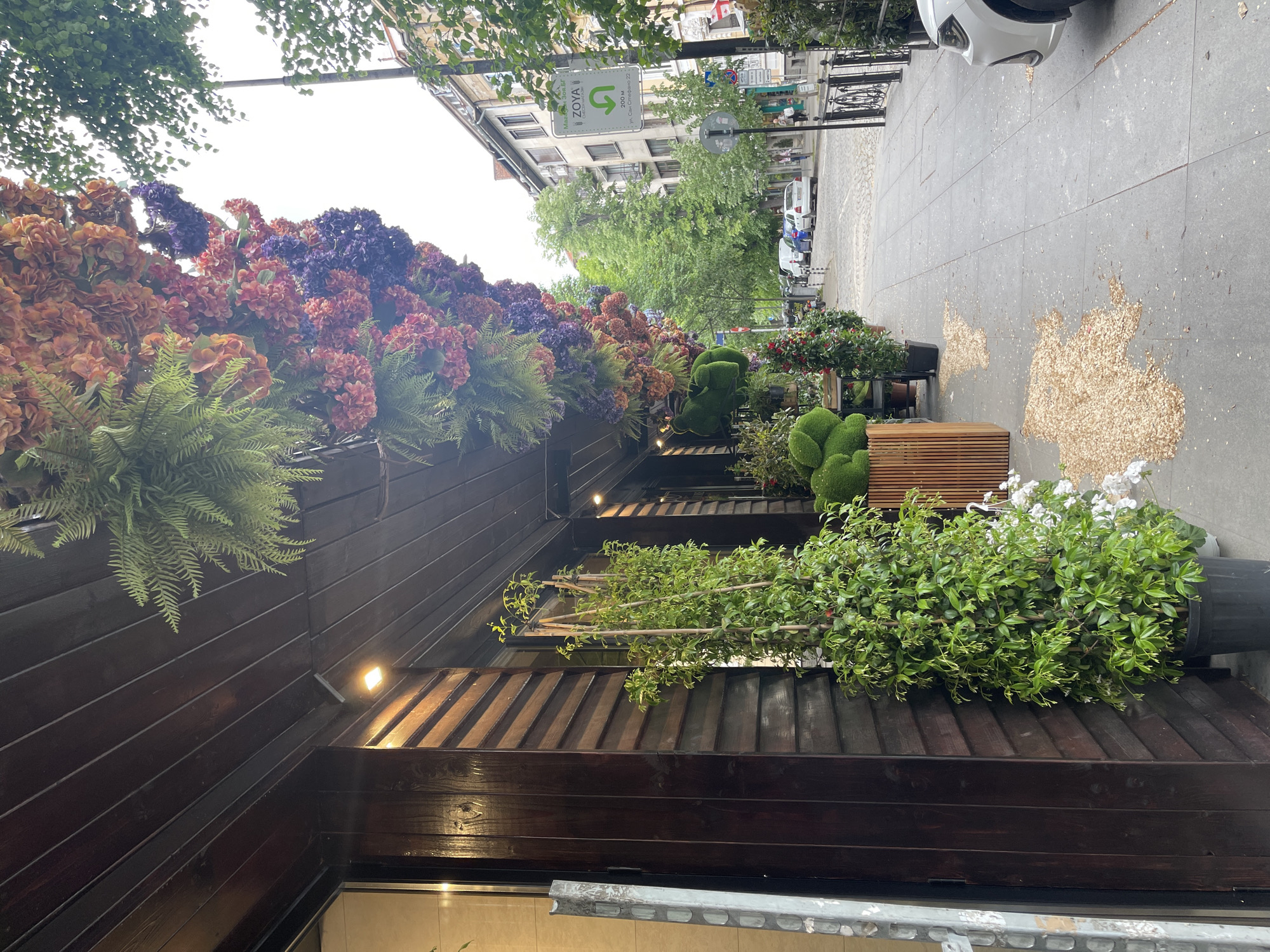 The image shows the exterior of a building adorned with vibrant floral decorations, located in the city of Sofia, Bulgaria. The building's facade is constructed with dark wood panels and features planter boxes overflowing with an assortment of artificial flowers in shades of orange, red, purple, and green ferns. Potted plants, including sculpted bushes, line the sidewalk, creating a lush green barrier. The sidewalk itself is gray and speckled with fallen leaves or other debris. In the background, a street scene unfolds with parked cars and pedestrians visible further down the road. A street sign with the word "Zoya" and Bulgarian text is also visible, indicating the location within Sofia. The lighting suggests an overcast day.