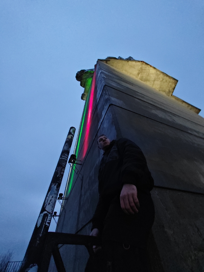 The scene depicts a low-angle view of a segment of the Monument to 1300 Years of Bulgaria in Shumen, Bulgaria, under twilight or nighttime conditions. A male individual, appearing to be a young adult, stands in the mid-ground, positioned slightly to the right of a tall metal pole. He is oriented facing upwards and slightly to his right, observing the monumental concrete structure. He wears a dark, possibly padded, jacket and dark trousers; his left hand hangs at his side. His face is partially illuminated by ambient light, while his body is predominantly in shadow.
The brutalist-style concrete monument dominates the upper and right portions of the frame, displaying angular forms and a textured surface. A prominent vertical strip of artificial lighting, featuring alternating red and green segments, runs along a visible edge of the monument's facade. Upper sections of the concrete structure are illuminated by a distinct yellowish light.
To the left of the individual, a tall, slender metal pole stands vertically, adorned with numerous stickers and some graffiti. Multiple small, dark-colored spotlights are mounted on this pole, directed towards the monument. A dark, angular metal structure, possibly a railing or support, is partially visible in the lower left corner. The sky above is a uniform, desaturated blue-gray.