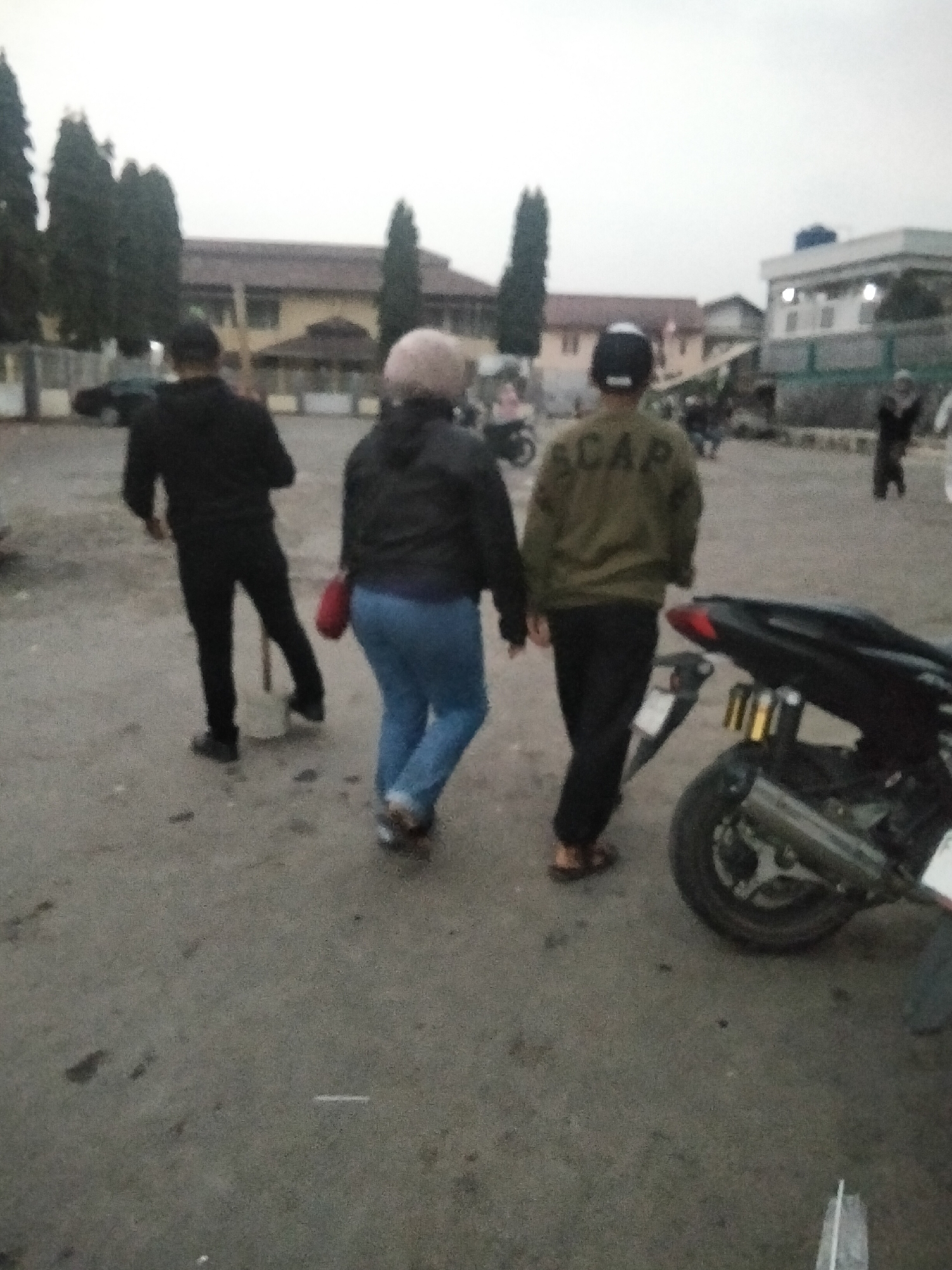 In the city of Cisaat, Indonesia, a low-angle, blurry photograph captures a scene on an outdoor paved area, possibly a parking lot or street, under an overcast sky. Three individuals are walking away from the camera. On the left, a person in a black long-sleeved shirt and black pants walks, holding a shovel with a long handle. In the center, a person wearing a light-colored head covering and a dark jacket walks with a red bag over their shoulder, clad in blue jeans. To the right, a person in a khaki jacket with "SCAP" written on the back in dark letters, and wearing a black baseball cap and dark pants, walks alongside the second individual. A dark-colored motorcycle with a prominent exhaust pipe is parked in the foreground on the right. In the background, there are buildings with tiled roofs and trees.