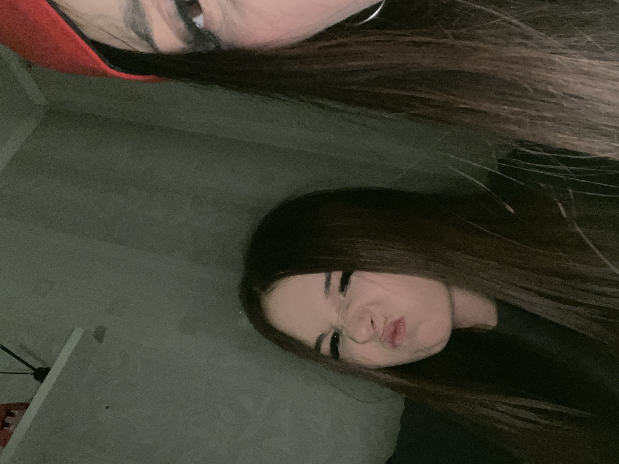 This image presents a close-up, tilted indoor shot featuring two individuals, likely captured in Saint Petersburg, Russia. The lighting is dim, suggesting an evening setting or a poorly lit room.
In the upper portion of the frame, the eye, eyebrow, and forehead of the first person are visible. They have long, dark hair and are wearing a red garment or accessory, possibly a hood. A silver hoop earring is discernible near their ear.
More centrally and prominently, the second person is depicted with their face tilted. They also have long, dark hair and are making a playful "duck face" expression, with squinted eyes and pursed lips. They appear to be wearing a dark top.
The background primarily consists of a plain, light greyish-green wall with a subtle texture. To the far left, a white, flat surface, possibly a shelf or part of a furniture item, is partially visible. Behind this, indistinct dark lines and a red shape resembling the letter "E" can be vaguely seen, hinting at a sign or decorative object.
The image conveys a casual and lighthearted mood, indicative of a spontaneous selfie or a fun moment shared between the two individuals.