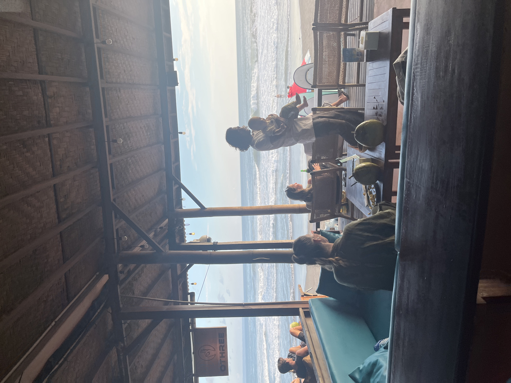 The image depicts a beachside cafe in North Kuta, Indonesia, during what appears to be late afternoon or early evening. The cafe has a thatched roof supported by wooden beams and columns, with open sides overlooking the ocean.
In the foreground, a person with long dark hair is carrying a baby. They are standing on the wooden deck of the cafe. To the left, a woman with blonde hair tied in a bun is seated at a table. In the mid-ground, several other patrons are visible, including a man reclining on a sofa and a woman sitting at a table facing away from the camera.
The beach and ocean are clearly visible in the background. Waves are breaking on the shore, and the sky is a clear blue with a few scattered clouds. Surfboards are propped up on the sand, suggesting a popular surfing spot.
Objects on the tables include drinks, coconuts, and possibly plates of food. The overall atmosphere seems relaxed and casual, consistent with a beach cafe setting. The lighting suggests a soft, natural light, likely from the setting sun.