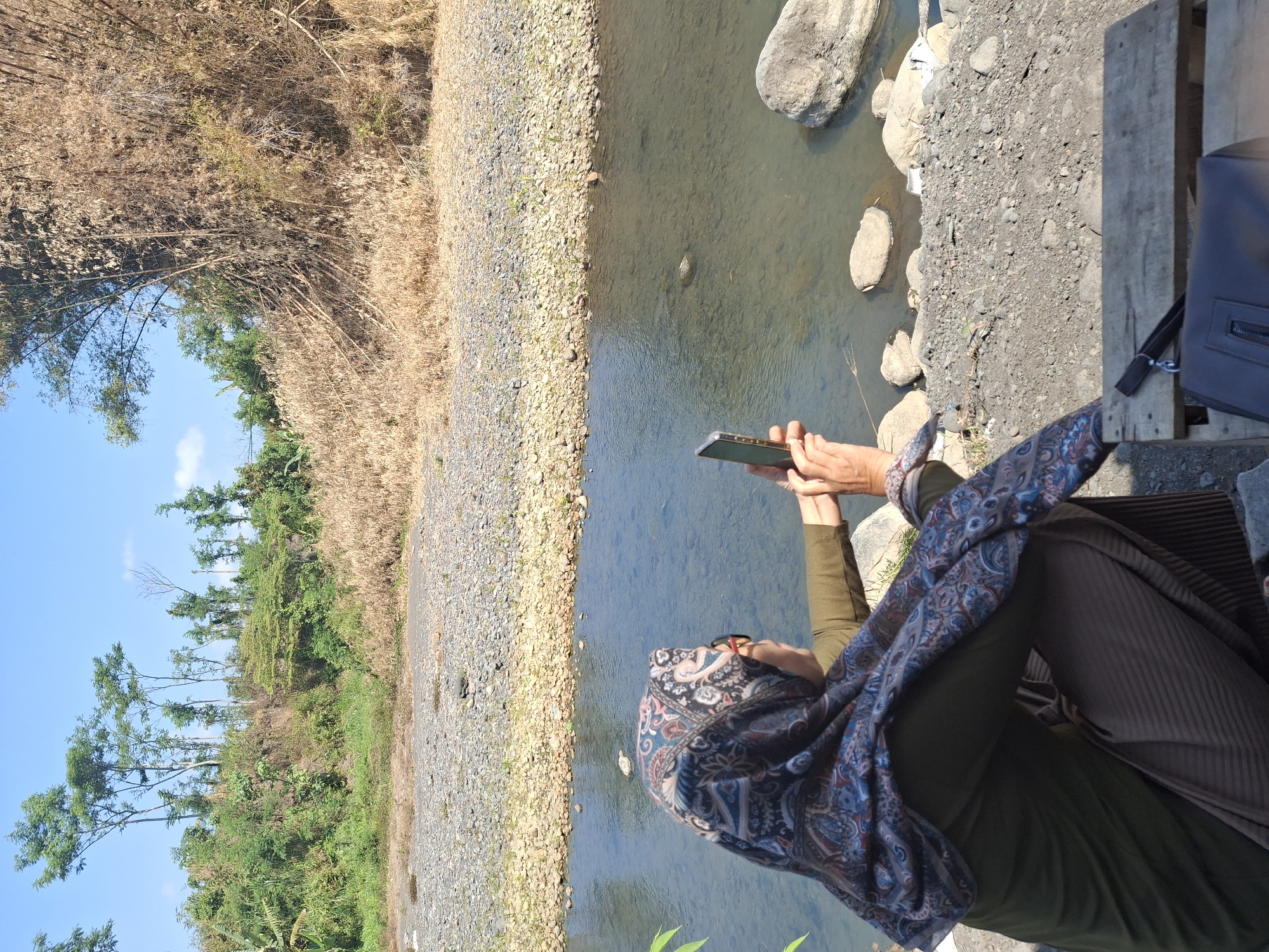 A woman wearing a hijab and glasses is squatting on the bank of a river in Lumajang, Indonesia, holding a smartphone and taking a photo. The river is clear and shallow, with a rocky bed visible. The surrounding environment is rural, with dry grasses and bamboo trees on the elevated bank opposite the river. The sky is clear and blue. The woman is dressed in a green long-sleeved shirt, brown striped pants, and a patterned hijab. She is holding the phone horizontally in her right hand. In the foreground, a wooden plank is visible, with a bag and a phone case on it. The scene appears to be a serene, natural landscape.