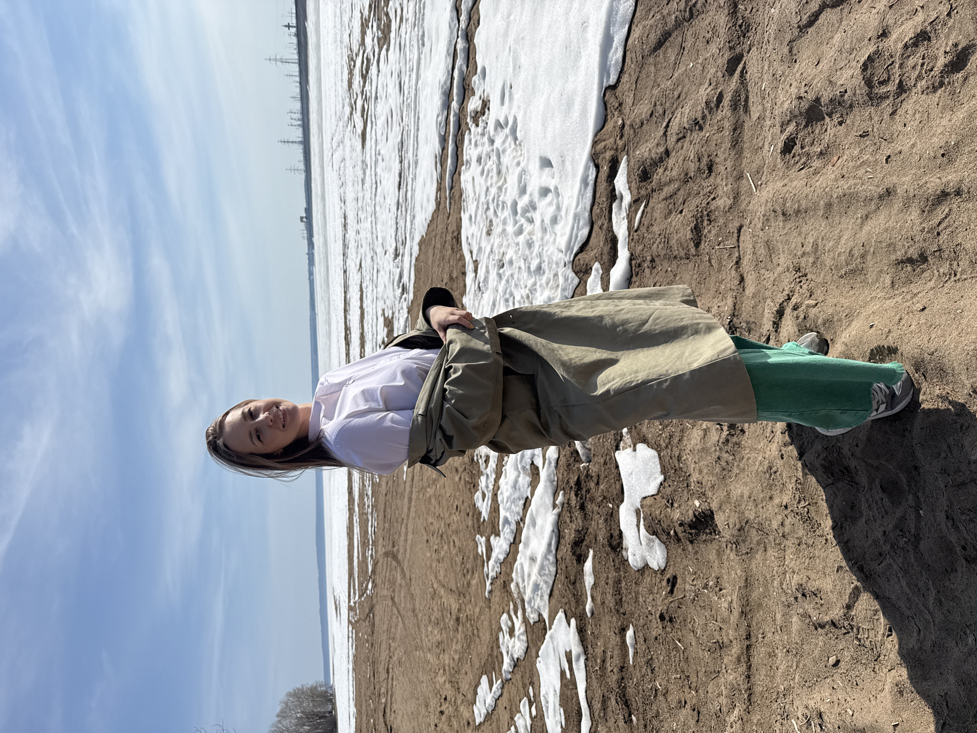A young woman is standing on a sandy beach with patches of snow in Noviy, Russia. She wears a white shirt, green pants, a khaki trench coat, and grey sneakers. The sky is blue with some clouds. In the distance, there is a body of water with snow cover and transmission towers.