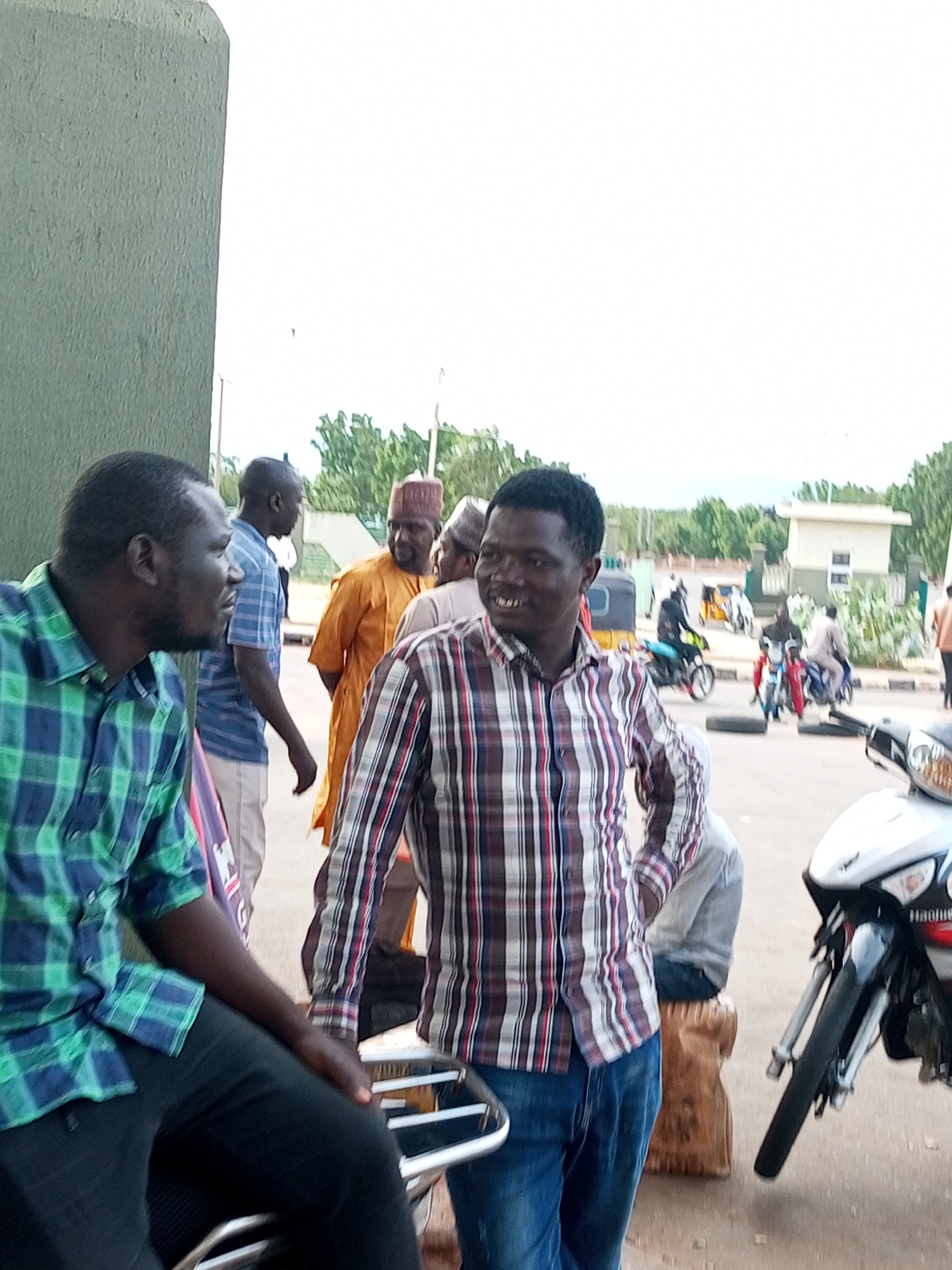 The image shows a group of people outdoors, likely in a street or open area, with a focus on a few individuals in the foreground. On the left, a man wearing a green and blue plaid shirt is seated, looking to his right. To his right, another man in a red, white, and black plaid shirt is standing and facing the camera, smiling. Behind them, several other people are visible. One man is wearing an orange traditional garment and a red cap. Another man, wearing a blue striped shirt, is in the background, as is a person in a light-colored garment.
In the background, there are trees and some buildings, including a light-colored structure with a dark roof. Several motorcycles are also present, with one prominently displayed in the right foreground, a white scooter-like motorcycle. Other motorcycles, some with riders, are visible further back, suggesting a busy or common gathering place, possibly a transportation hub or market area. The scene appears to be outdoors during the day, with bright, overcast lighting. The overall mood of the people in the foreground seems relaxed and friendly, especially the man in the plaid shirt who is smiling at the camera. There is no discernible text visible in the image.