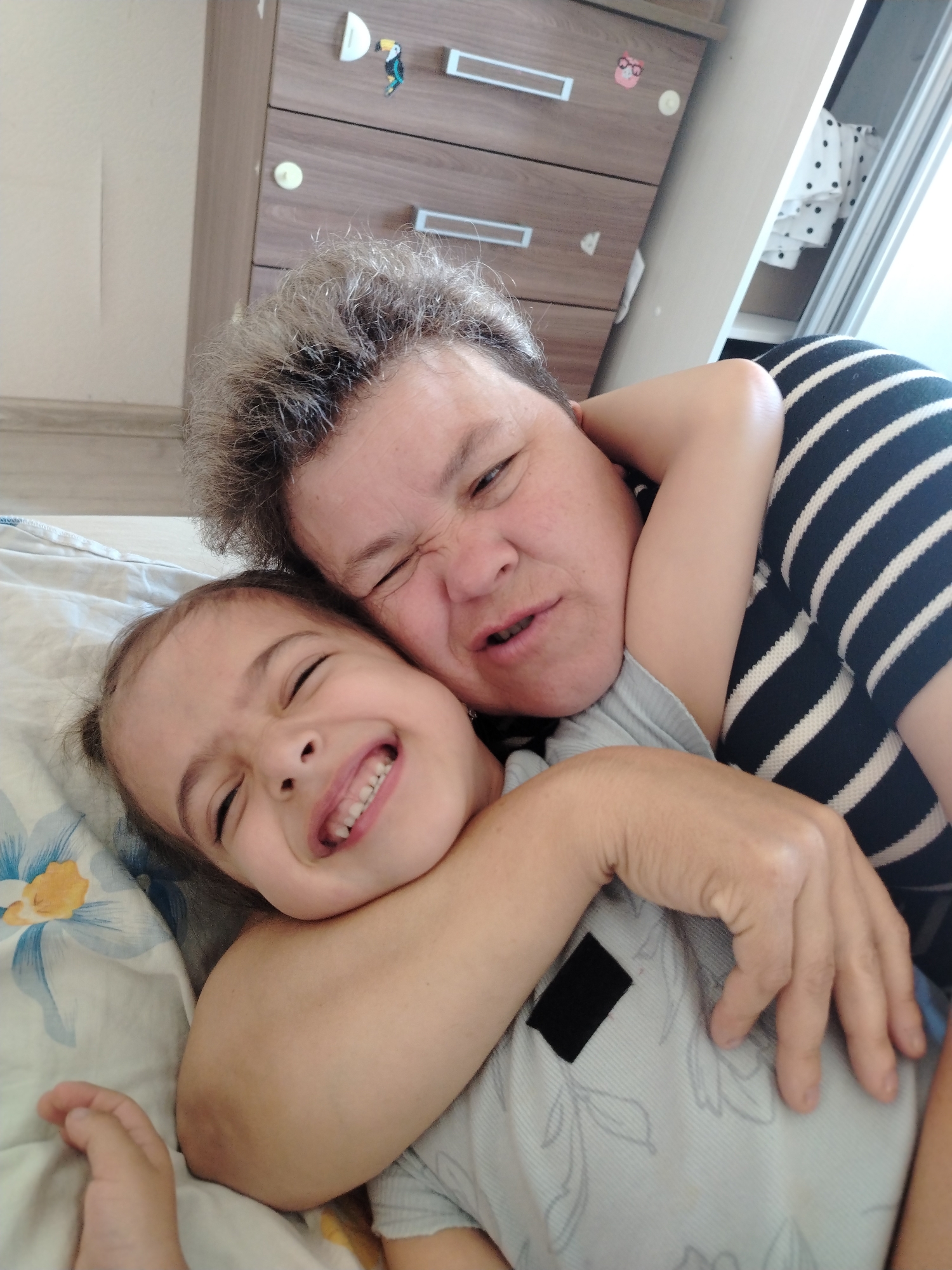 The image shows two individuals, an adult and a child, embracing on a bed. The adult, with short gray hair, is wearing a black and white striped shirt. The child, with dark hair, is wearing a light blue shirt with a subtle pattern. They are both smiling with their eyes closed. The background includes a wooden dresser with several drawers and some decorative items. A window with a white curtain is visible to the right. The setting appears to be an indoor bedroom. There is no information available about the specific location in the city of Noviy, Russia.