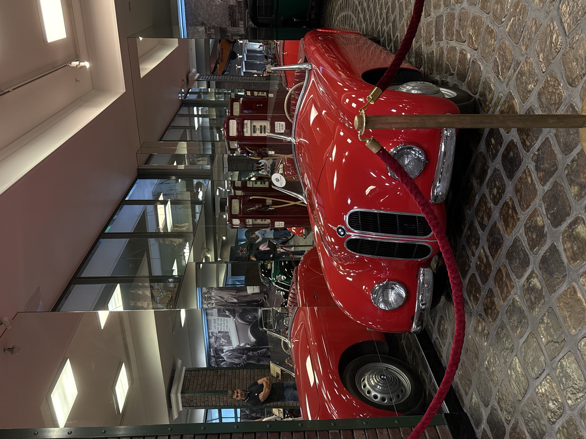 The image shows the interior of a museum with several classic cars on display. In the foreground, a bright red convertible with a black grille and chrome accents is prominently featured. The car has a tan interior and its windshield is partially raised. To the left of this car, another red convertible is partially visible, showing its driver's side door, fender, and wheel. Behind the red cars, vintage gas pumps are visible, with "CHEVRON" branding on one of them. In the background, there are large mirrors reflecting the scene and the ceiling, which is illuminated by fluorescent lights and track lighting. Several people are present, including a man standing to the left of the foreground car, arms crossed, looking towards the camera. Other individuals are seen in the reflection and further back in the museum. The floor is paved with cobblestone-like tiles. A red velvet rope on stanchions is present in the foreground, separating the viewer from the exhibit. The overall environment appears to be a well-lit museum hall dedicated to vintage automobiles. This scene is located at the Arkhangelskoe Museum in Arkhangelskoye, Russia.
