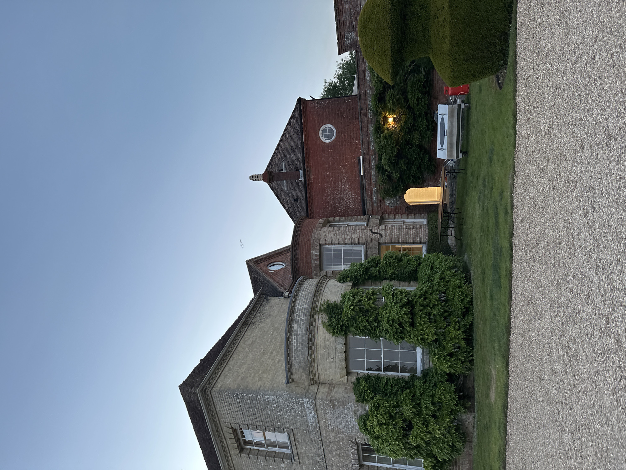 Exterior shot of a building with greenery in an unknown location. The building is made of brick and has several windows. There is ivy growing on the side of the building. There are also some trees and bushes in the yard. The sky is clear.