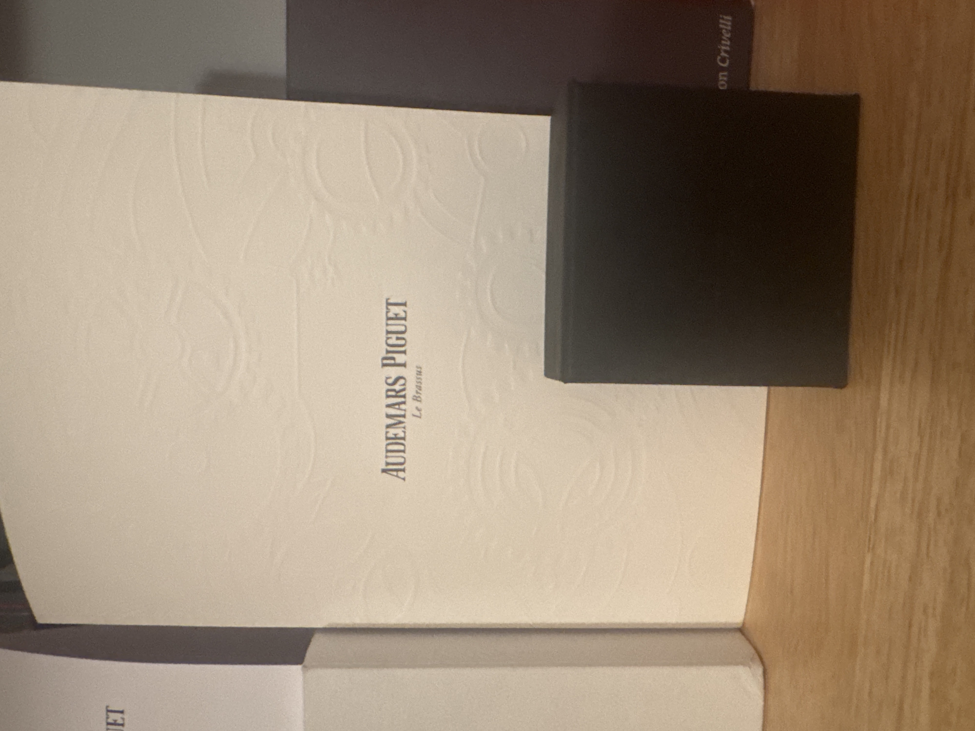 The image displays an indoor still life arrangement, rotated 90 degrees clockwise. The main subject is a large, light-colored (off-white or cream) document folder or book, positioned centrally and appearing to be open or partially open. This item features a subtle, embossed pattern, possibly an abstract or floral design, across its surface. Prominently displayed vertically on this item is the text "AUDEMARS PIGUET" in a dark font, with "Le Brassus" written in smaller text directly below it.
Resting on the right side of the light-colored item is a small, rectangular black box. In the background, a dark-colored book or folder is partially visible behind the main document holder. Text partially visible on this background item reads "on Crivelli". Another light-colored item with partial text "ET" is visible at the very bottom left.
The setting appears to be an interior space, likely a table or desk with a wooden surface visible on the right side and partly at the bottom. The lighting suggests ambient indoor illumination. The scene depicts luxury brand items from Audemars Piguet, likely within an indoor setting in Milan, Italy. No people are present, and no specific activity is taking place beyond the arrangement of these objects.