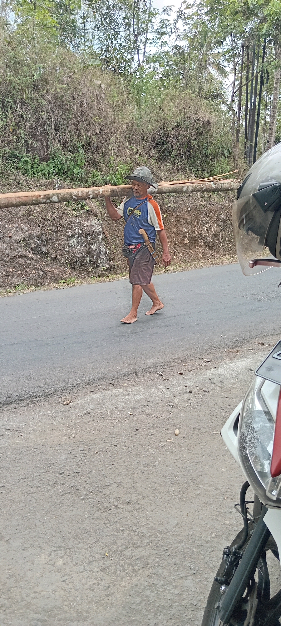 An elderly man walks barefoot along a paved road, carrying a long, thick log horizontally on his right shoulder. He is wearing a grey, patterned hat, a blue t-shirt with orange sleeves, and brown shorts. A knife with a curved blade is sheathed at his waist. The road is flanked by a dry, grassy embankment on the left, with sparse trees and vegetation visible further up the slope. On the right side of the image, the front of a white motorcycle with a clear windshield is partially visible, suggesting the photograph was taken from another vehicle. The sky is overcast. The location is Deudeul, Indonesia.