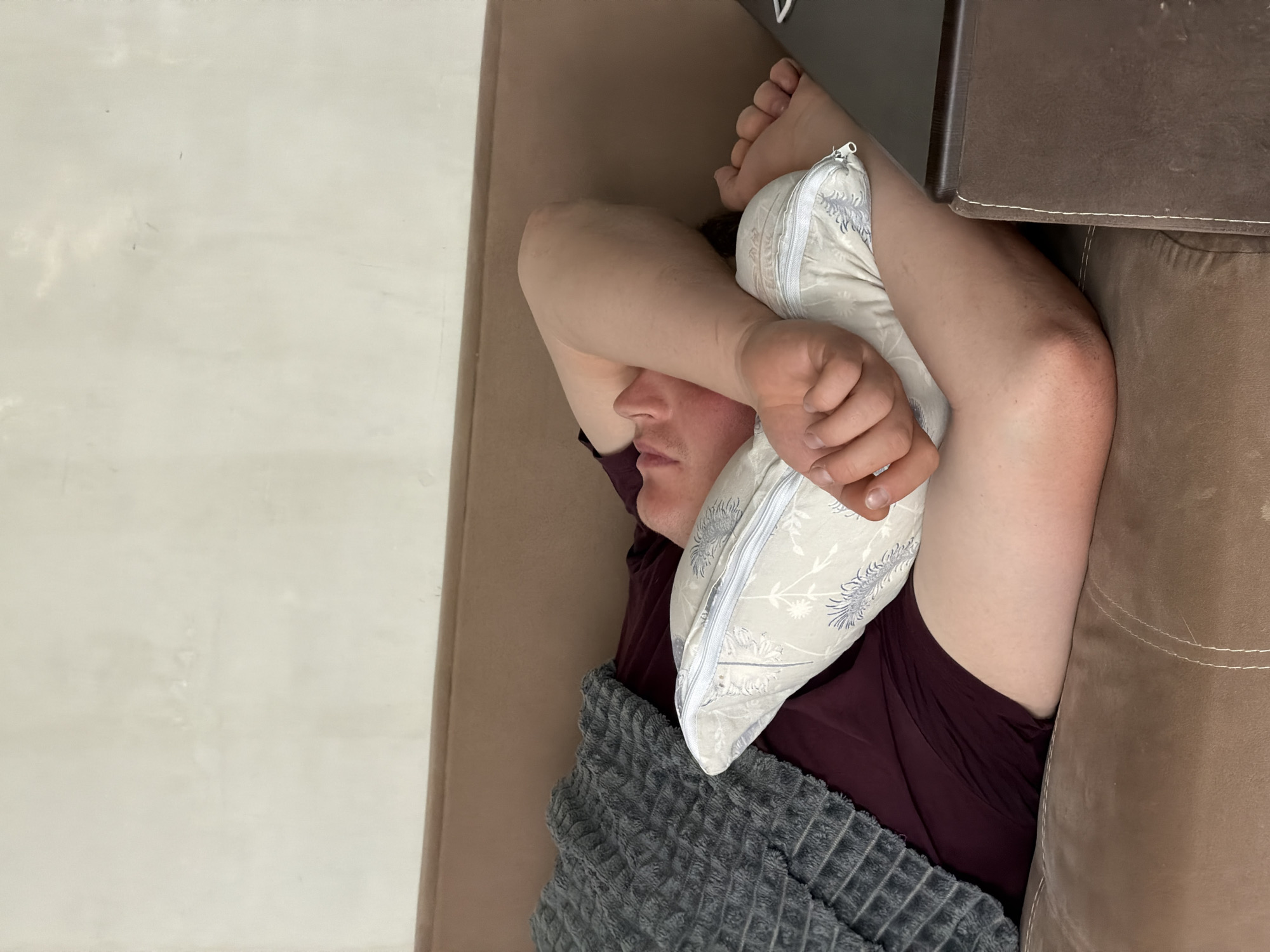 A man lies on a tan couch, shielding his face with a pillow and an arm. He is partially covered by a gray blanket and wears a maroon shirt. A dark brown table is positioned next to the couch. The location is Chaikovsky, Russia.