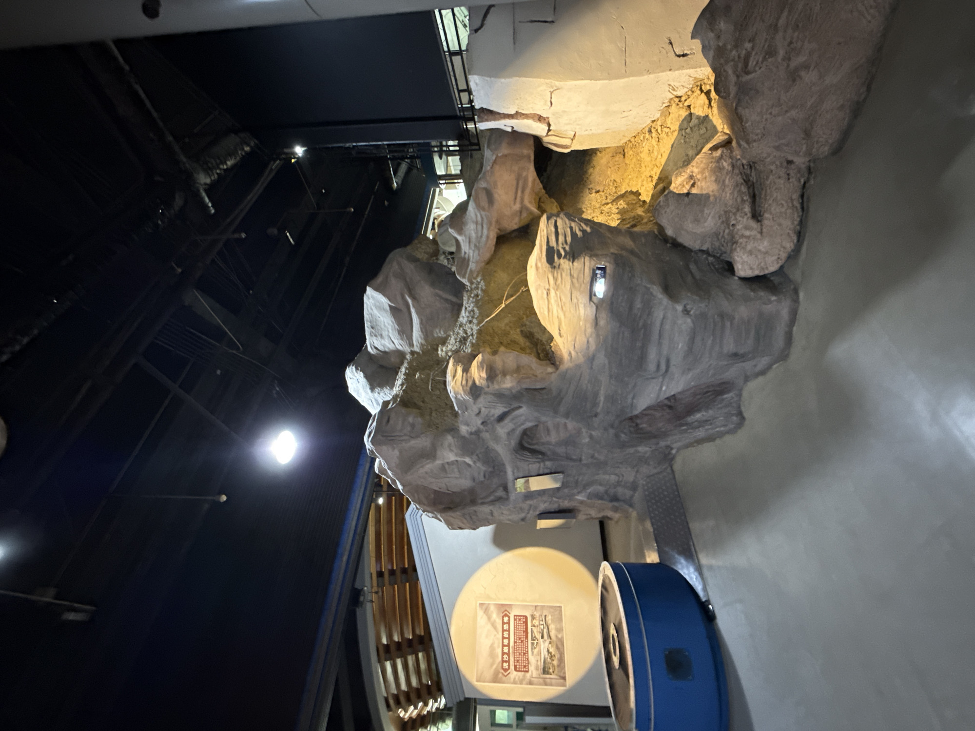 The image shows an interior space in 江北城街道, China, featuring a large, artificial rock formation illuminated by warm lighting. The rock structure appears to be part of an exhibit or display, potentially in a museum or public space. Below the rock, there is a white circular display with Chinese text and images, positioned next to a blue cylindrical object. The background includes dark, industrial-looking ceiling structures and support beams. The overall setting appears to be a modern exhibition or museum environment.