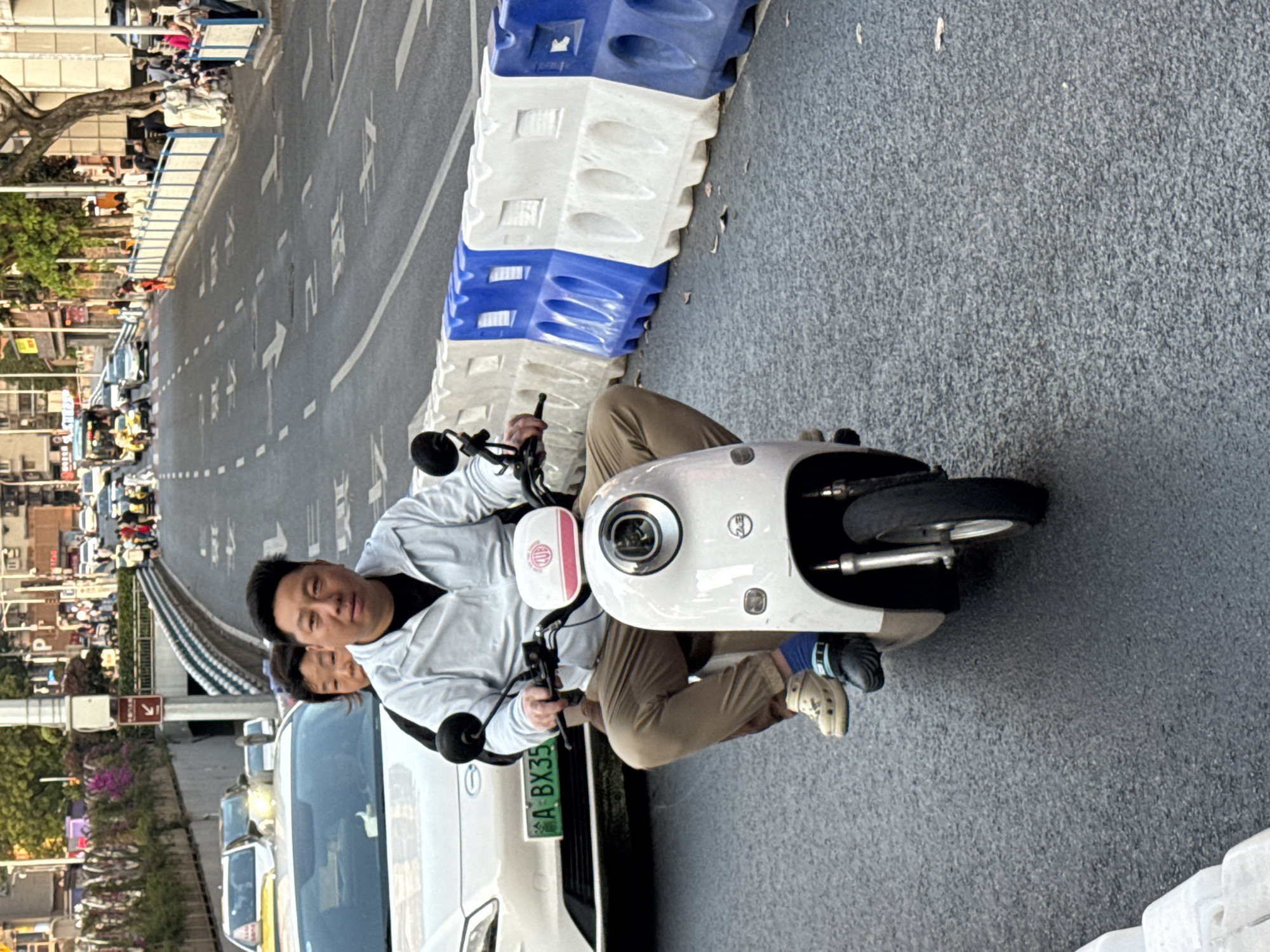 A medium-angle shot depicts two people riding a white electric scooter on a street in Yuzhong District, China. The rider is an Asian man in a light-colored jacket and tan pants, looking directly at the camera. A second person is seated behind him, partially obscured. The scooter is beside a white car, with a green license plate visible with the numbers and letters "A BX35". The street is paved and lined with white and blue barriers. In the background, there is a bridge and a street with cars and pedestrians. Buildings and trees are also visible in the distance. The overall lighting suggests it is daytime.
